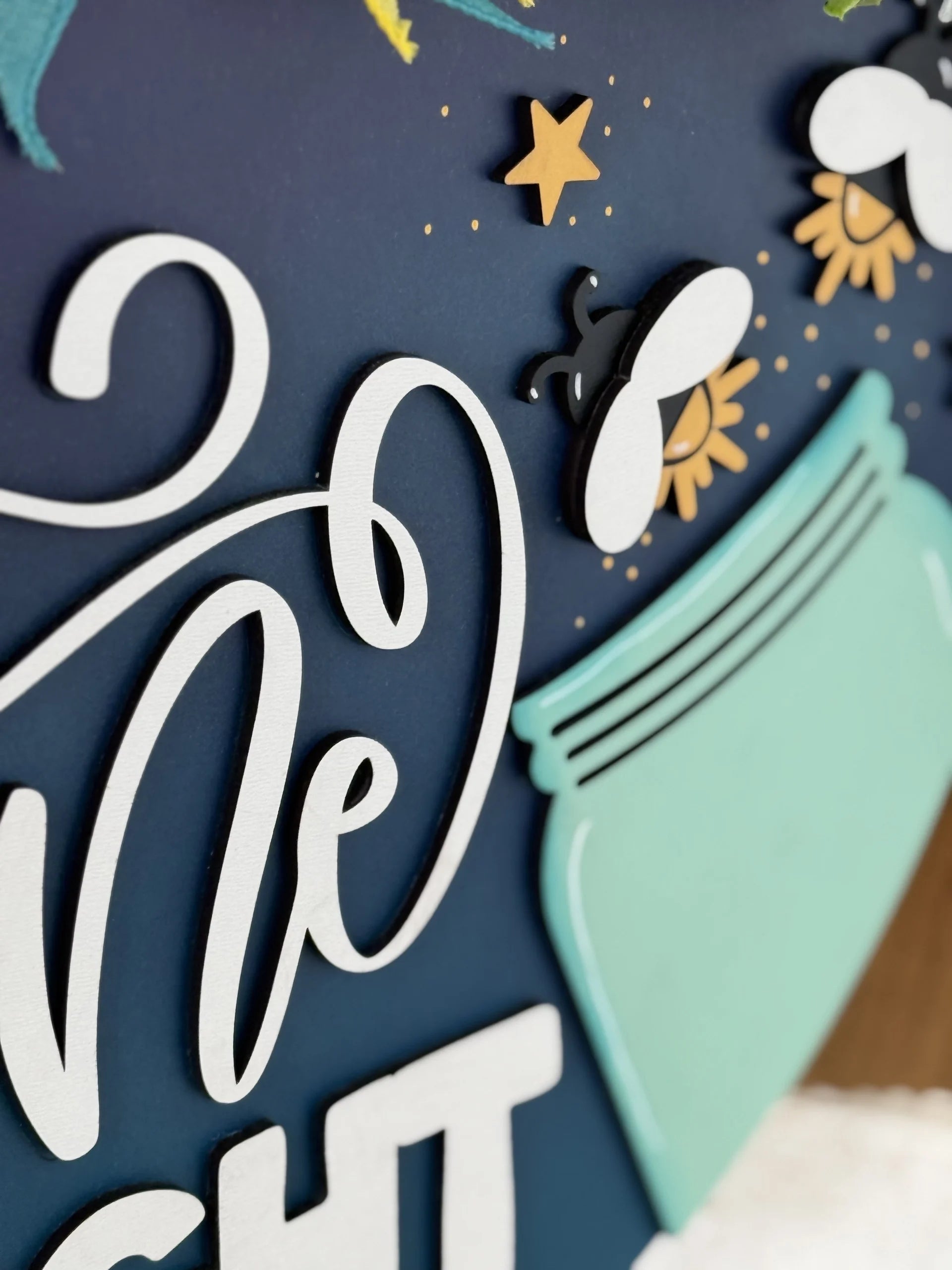 A close-up of a decorative sign on a dark blue background features white cursive text, yellow stars, illustrated fireflies with glowing tails, and part of a light blue jar, giving a whimsical, nighttime feel.