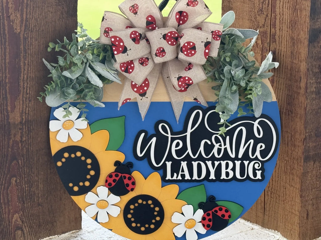 A round wooden sign features a large bow with red and black ladybug designs, leafy greenery, sunflowers, white daisies, and cartoon ladybugs. The sign has a blue bottom half and says “Welcome LADYBUG” in bold white and black letters.