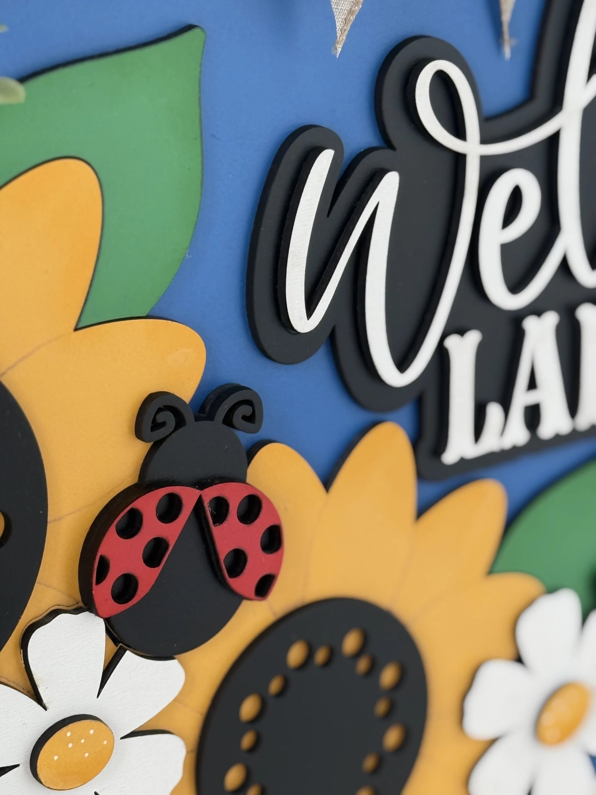 A close-up of a colorful decorative sign featuring large yellow sunflowers, white flowers, a red and black ladybug, and the word welcome in bold script letters on a vibrant blue background.