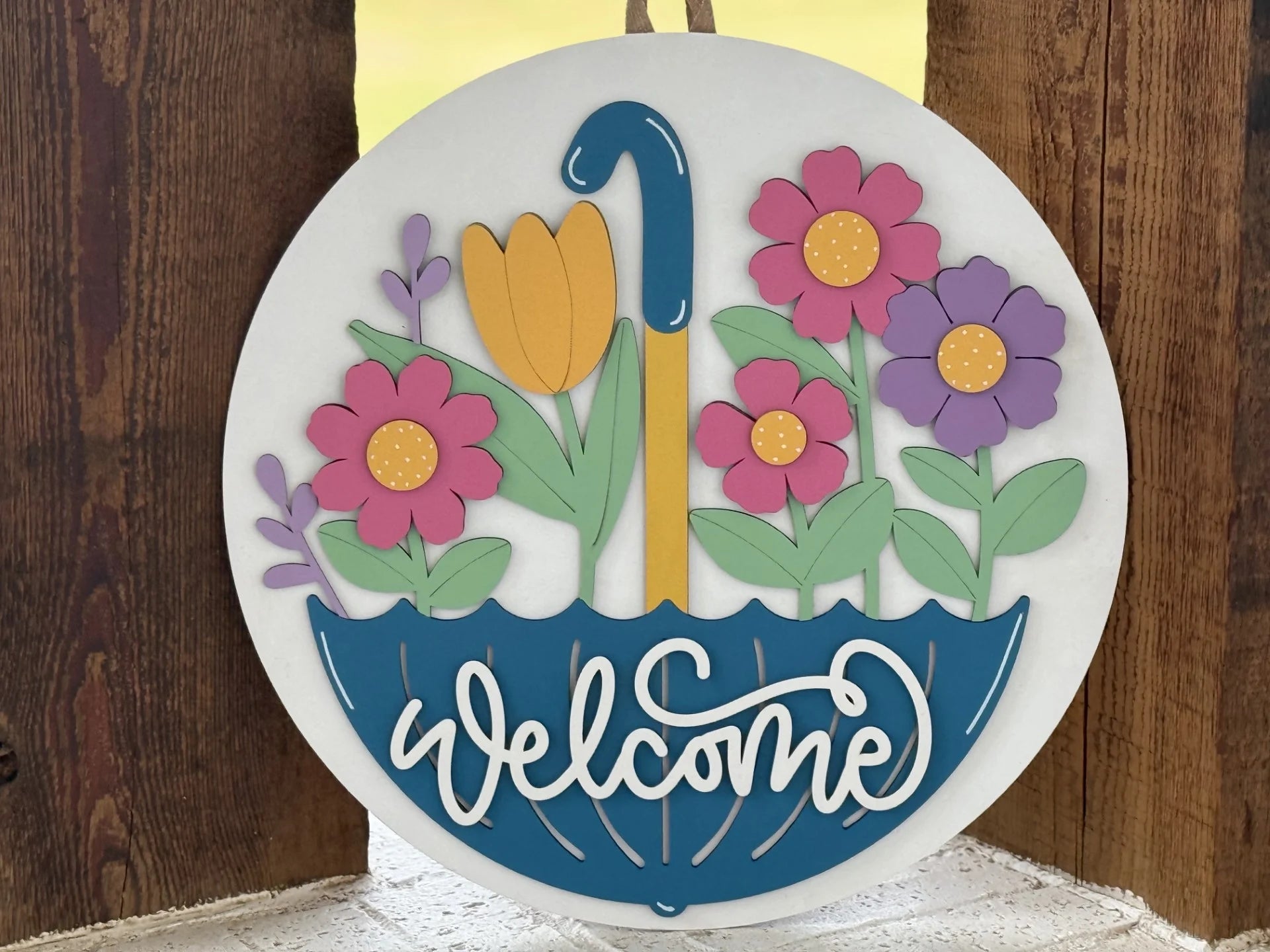 A round sign shows a blue umbrella filled with colorful flowers—pink, purple, and yellow—with green leaves. The word Welcome is written in white script on the umbrella. The sign hangs against a wooden background.