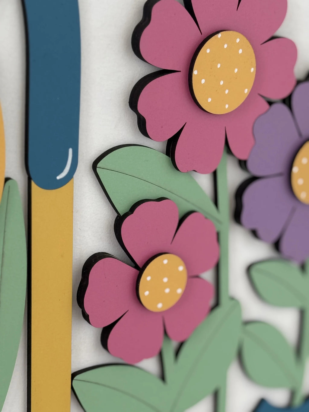 Close-up of a colorful, layered paper art piece showing pink and purple flowers with yellow centers, green leaves, and a blue-and-yellow vertical shape, all against a light background. The flowers and leaves have a cartoon-like, playful design.