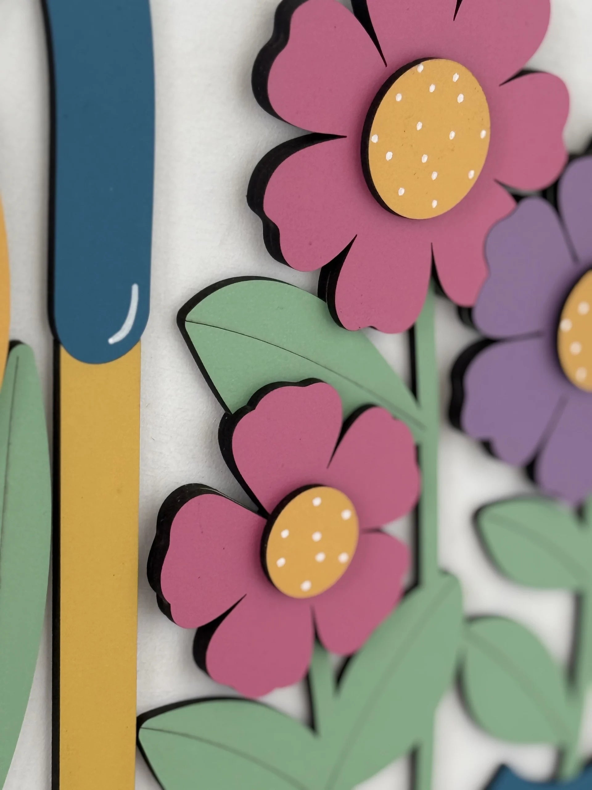 Close-up of a colorful, layered paper art piece showing pink and purple flowers with yellow centers, green leaves, and a blue-and-yellow vertical shape, all against a light background. The flowers and leaves have a cartoon-like, playful design.