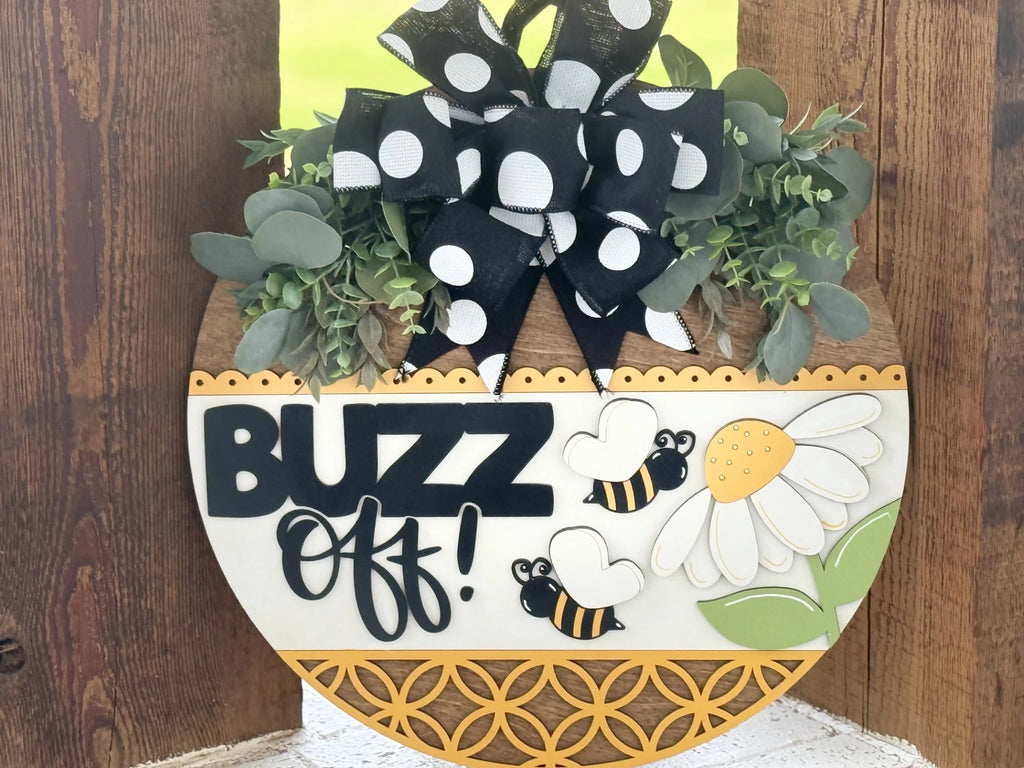 A round wooden sign decorated with greenery, a large black-and-white polka dot bow, a white daisy, two cartoon bees, and the words “BUZZ Off!” in bold black letters. The bottom edge has a yellow geometric pattern. The background is wood.