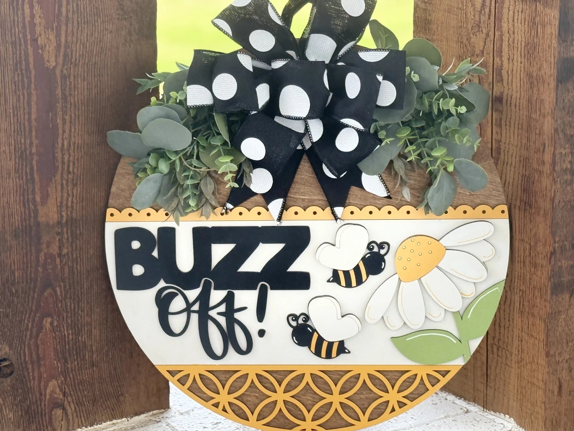 A round wooden sign decorated with greenery, a large black-and-white polka dot bow, a white daisy, two cartoon bees, and the words “BUZZ Off!” in bold black letters. The bottom edge has a yellow geometric pattern. The background is wood.