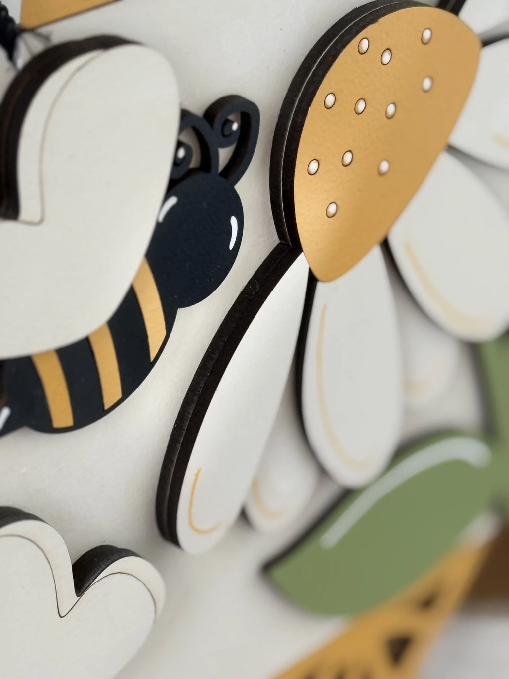 A close-up of a decorative piece featuring a cartoon-like bee with black and yellow stripes near a white and yellow daisy flower. The design is made from layered wood, creating a raised, 3D effect. Soft shadows and blurred background details are visible.