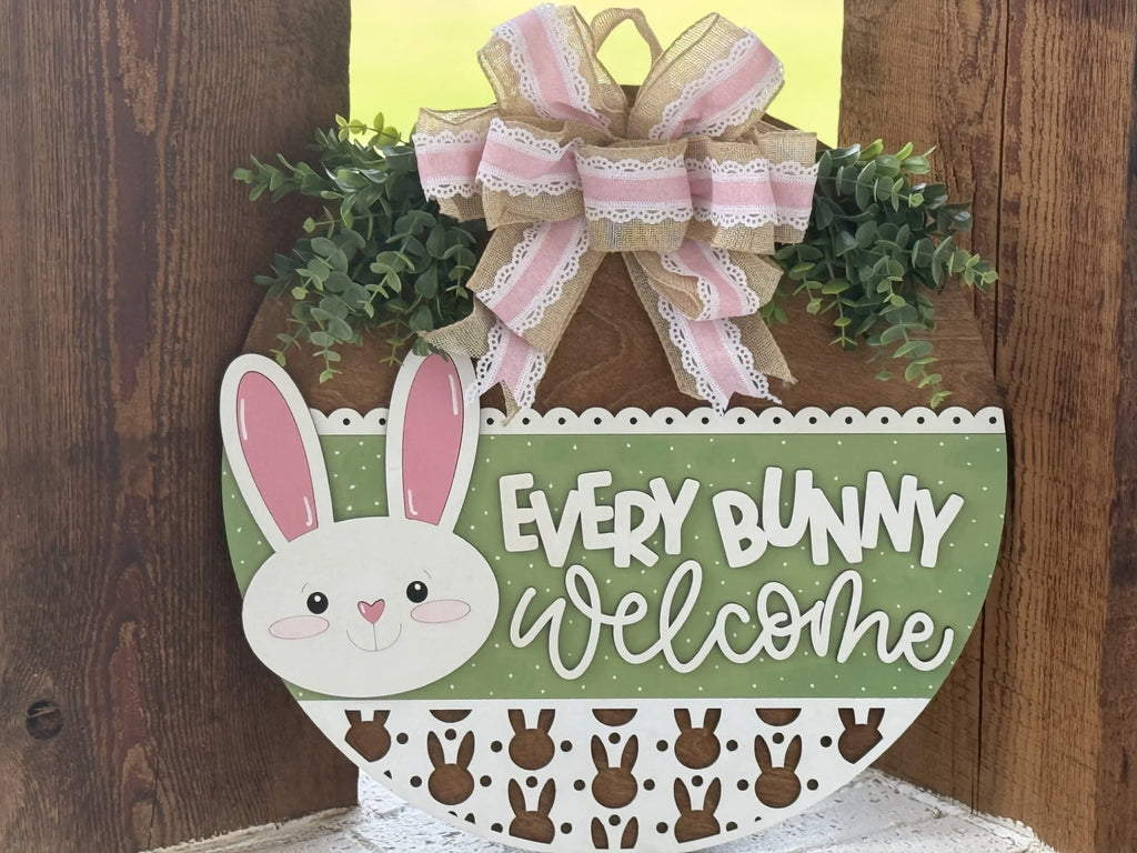 A wooden sign shaped like an Easter egg features a smiling bunny face, green polka-dot background, and the words “Every Bunny Welcome.” It’s decorated with a pink and lace bow, faux greenery, and a border of small brown bunny silhouettes.