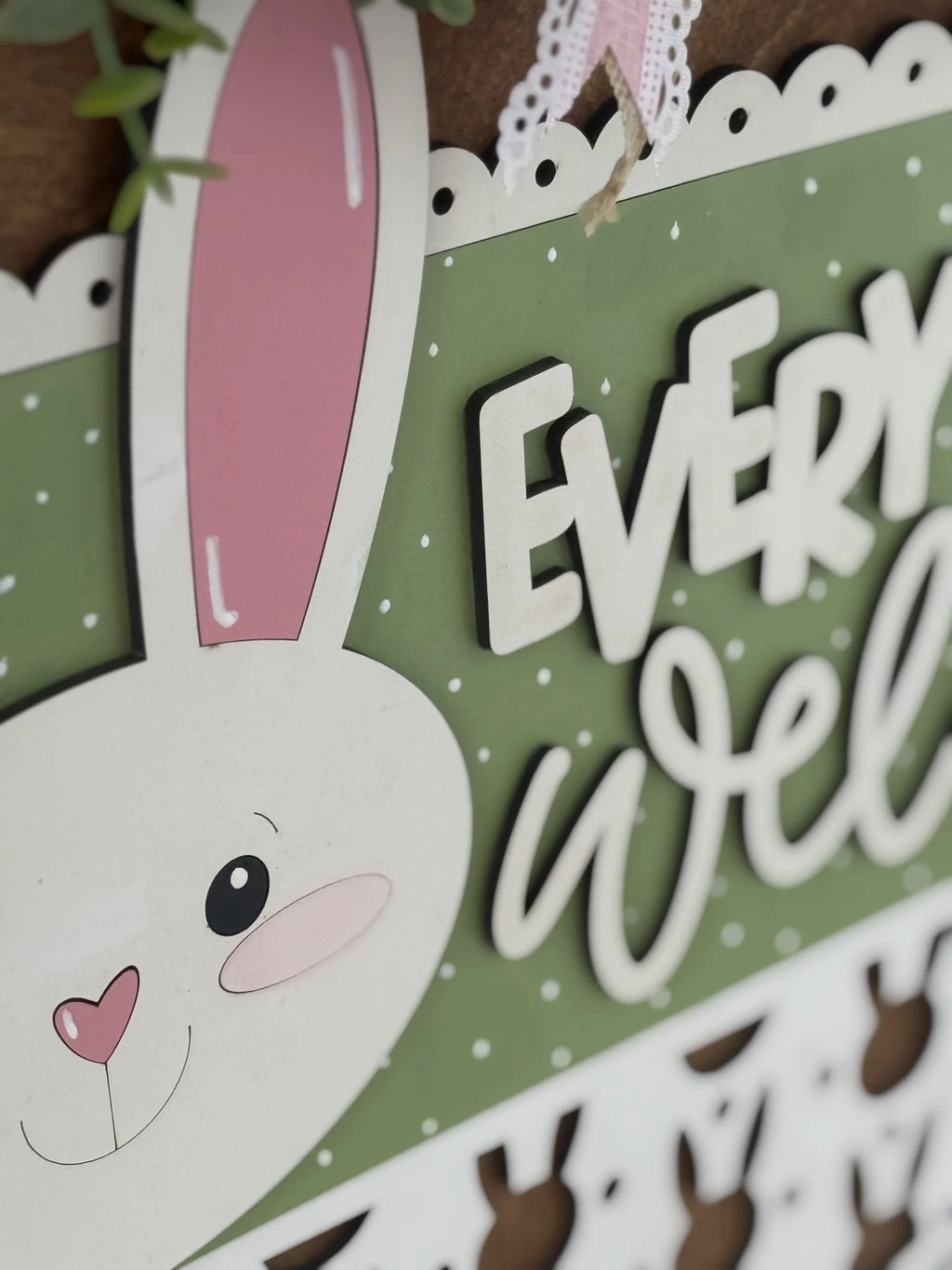 A close-up of a decorative sign featuring a smiling white bunny with a pink nose and ears. The background is green with white polka dots, and the visible partial text reads EVERY WE. The sign has scalloped edges and a lace ribbon at the top.