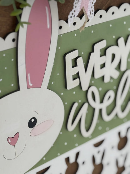 A close-up of a decorative sign featuring a smiling white bunny with a pink nose and ears. The background is green with white polka dots, and the visible partial text reads EVERY WE. The sign has scalloped edges and a lace ribbon at the top.
