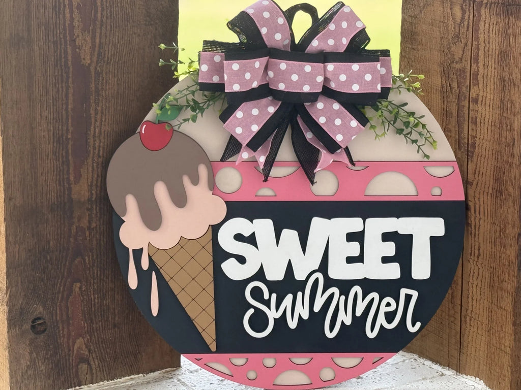 A round wooden sign features a pink and brown ice cream cone with a cherry, a large pink polka-dot bow with greenery, and the words SWEET summer in bold white letters on a black background. The sign has pink, brown, and black decorative elements.