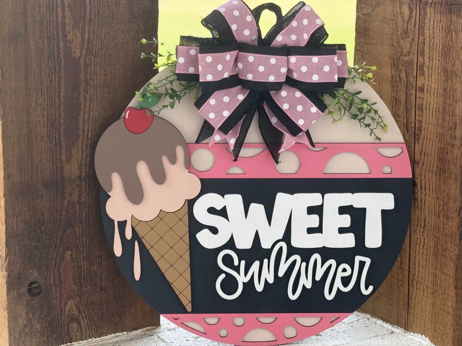 A round wooden sign features a pink and brown ice cream cone with a cherry, a large pink polka-dot bow with greenery, and the words SWEET summer in bold white letters on a black background. The sign has pink, brown, and black decorative elements.