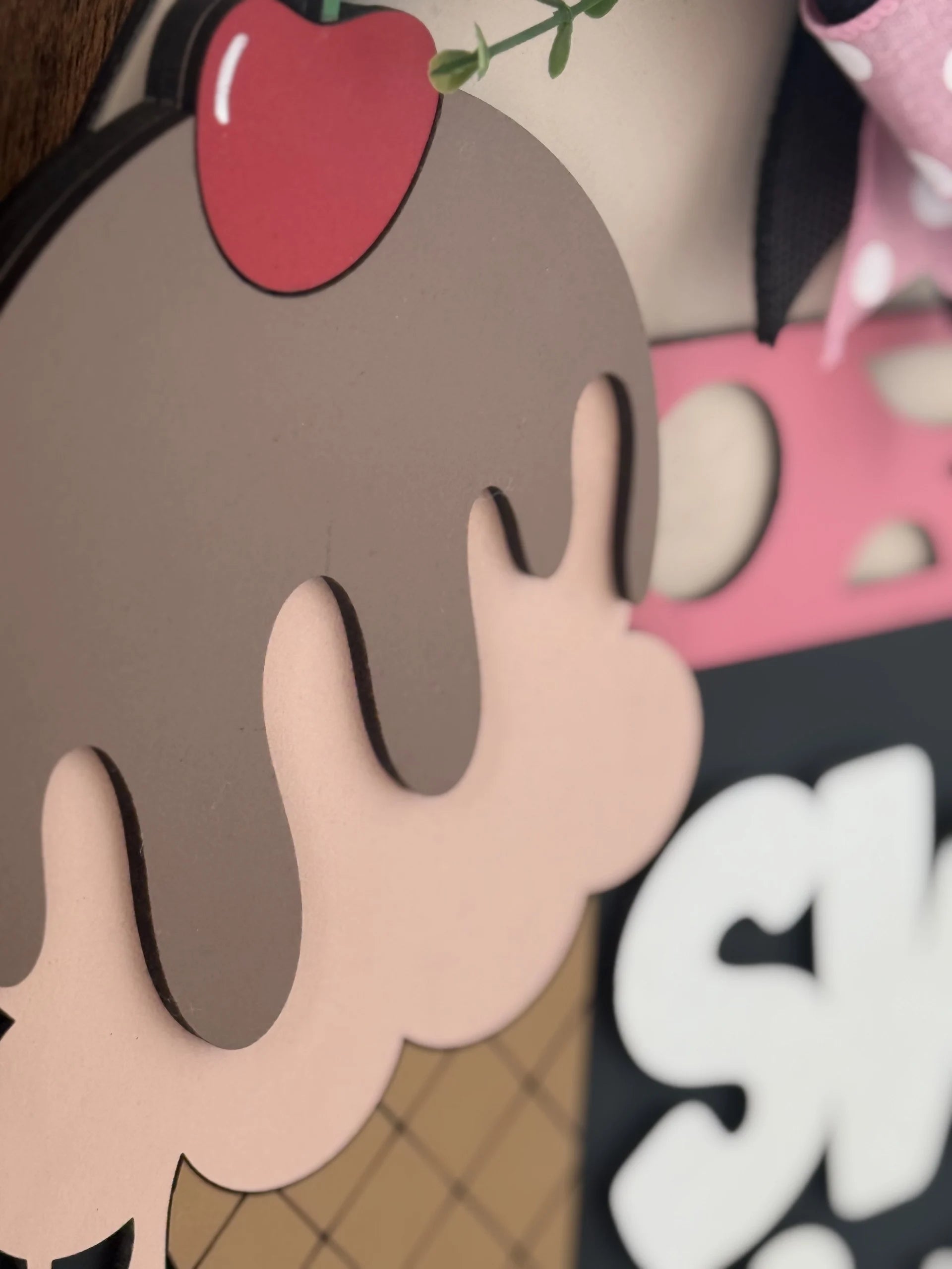 A close-up of a decorative ice cream cone illustration shows a pink cone with brown chocolate drips, a red cherry on top, and part of a sign with bold white letters on a black background, along with some pink and polka dot patterns in the background.
