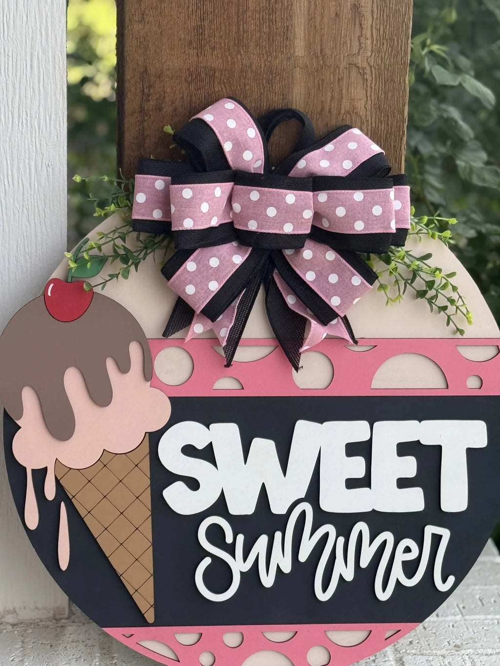 A decorative sign shaped like an ice cream cone with a cherry on top reads “SWEET Summer” in bold white letters. It features a large pink and white polka-dot bow with greenery arranged above the sign, and is placed against a wooden post.