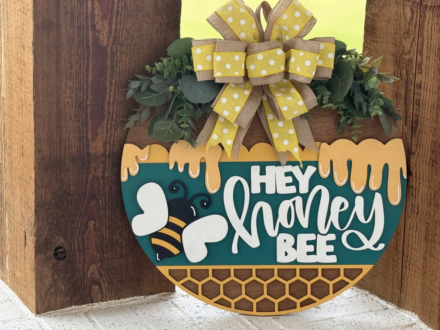 A round sign with yellow honeycomb and dripping honey designs says Hey honey bee in white script. A cartoon bee is pictured beside the text. The sign is topped with green leaves and a large yellow polka-dot ribbon bow.