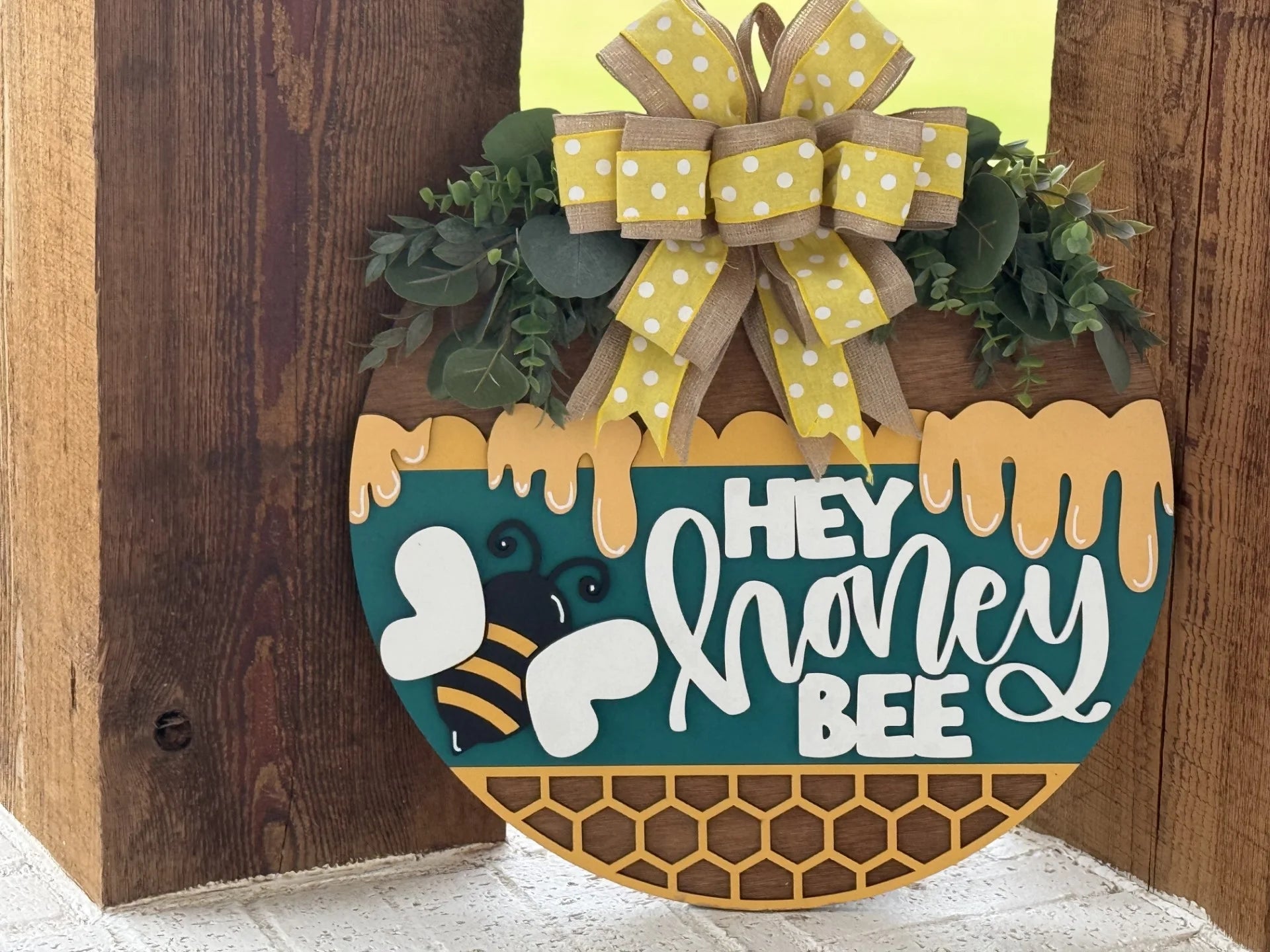A round sign with yellow honeycomb and dripping honey designs says Hey honey bee in white script. A cartoon bee is pictured beside the text. The sign is topped with green leaves and a large yellow polka-dot ribbon bow.