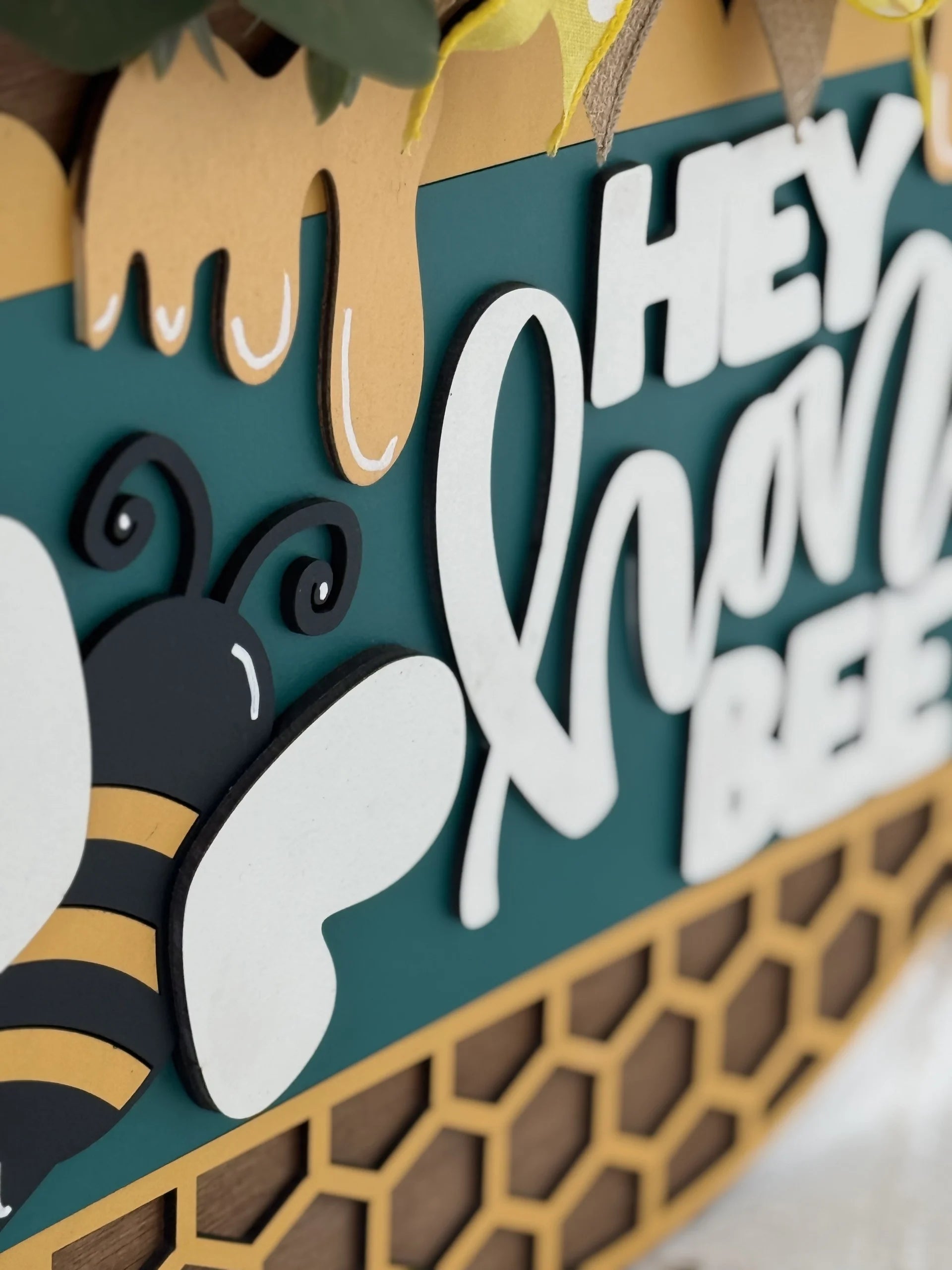 A close-up of a decorative sign features a cartoon bee with white wings and black-and-yellow stripes, honeycomb patterns, and the raised text HEY HONEY BEE in bold white letters on a teal background, with honey dripping from the top edge.