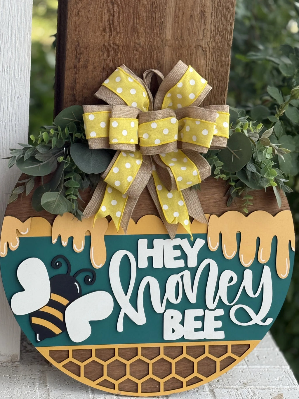 A round wooden sign hangs on a post, decorated with greenery and a yellow polka dot bow. The sign features honeycomb and honey designs, a cartoon bee, and the words “Hey Honey Bee” in playful white lettering.