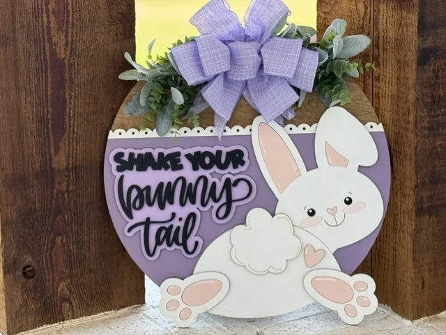 A round wooden sign features a smiling white bunny with pink ears and heart-shaped paw pads, a fluffy tail, and a lavender bow above its head. Text on a purple background reads, “Shake your bunny tail.” Greenery decorates the top of the sign.