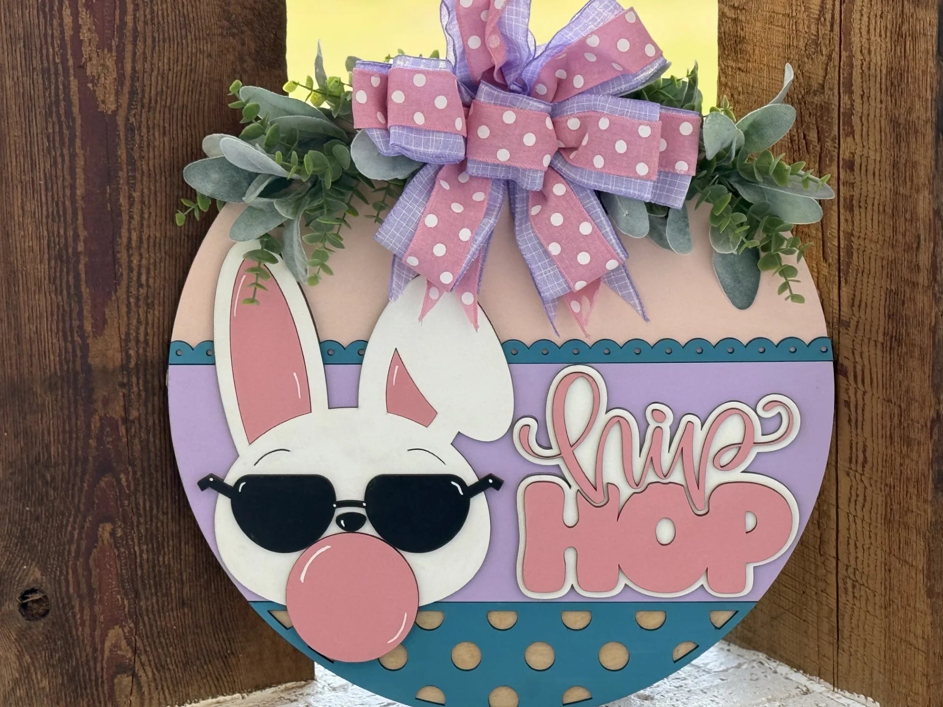 A round Easter sign features a bunny with sunglasses and a pink bubble gum bubble. The sign reads hip hop in large pink letters, and is decorated with polka dots, leaves, and a large purple polka dot bow at the top. The background is wood.