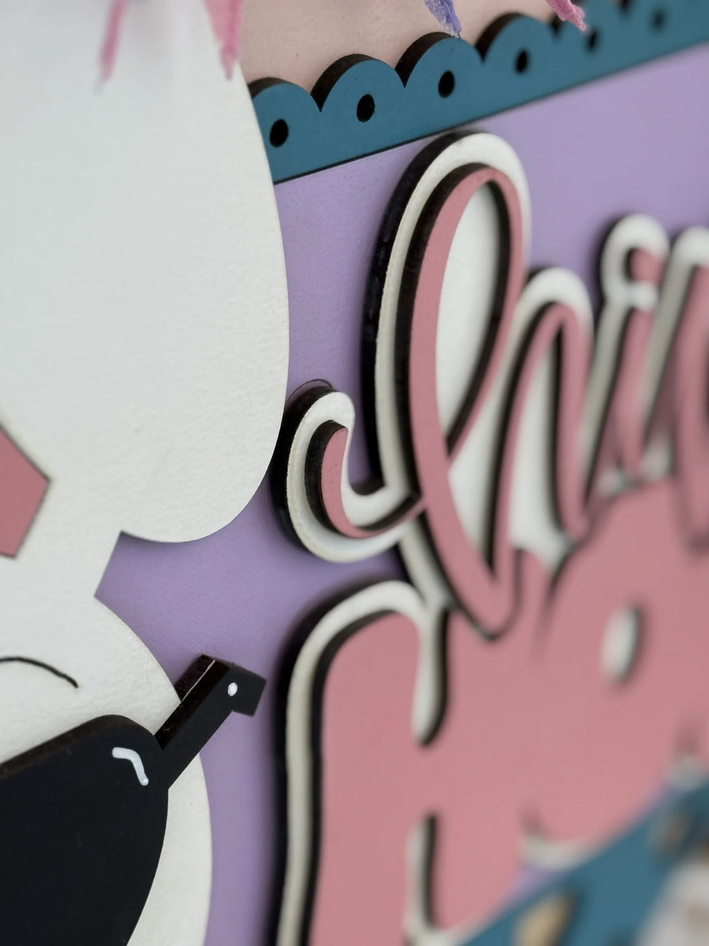 Close-up of a decorative sign with layered, cut-out lettering in white, pink, and black, set against a lilac background. Part of a white cartoon figure with a black wing and pink accents is visible on the left, with scalloped blue edging on top.