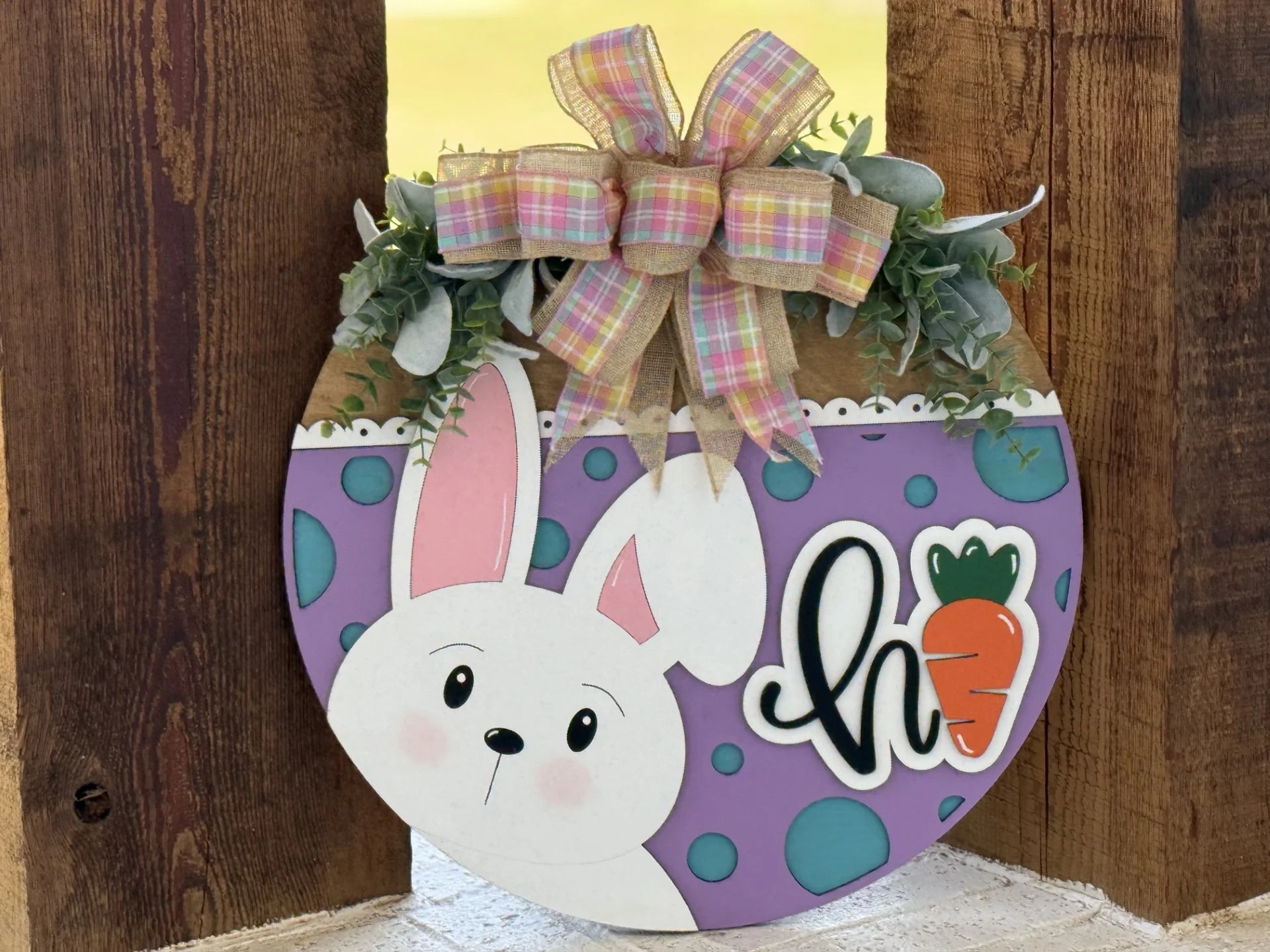 A round Easter sign features a cute white bunny with pink ears, a carrot, and the word hi on a purple background with teal dots. It’s topped with a plaid ribbon bow, greenery, and is set against a wooden backdrop.