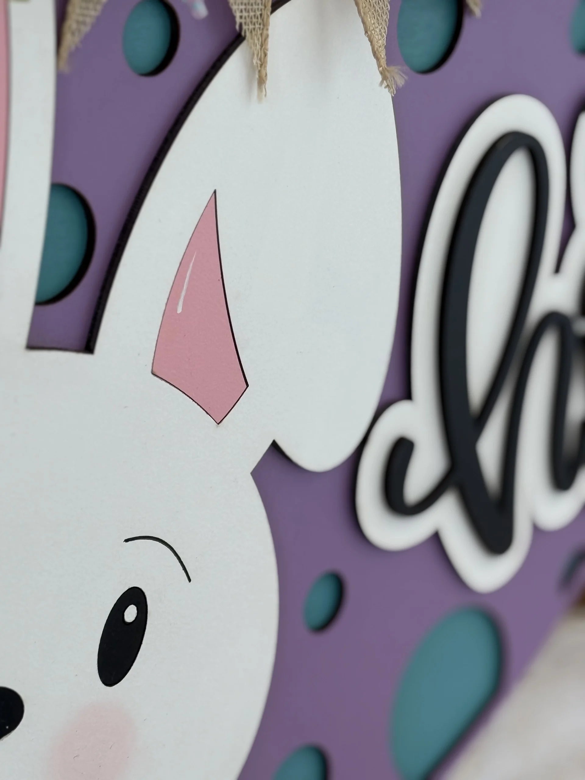 A close-up of a decorative sign features part of a white cartoon bunny with a pink inner ear, black eyes, and a hint of a smile, against a purple background with blue dots and partial cursive text in black and white. There is burlap fabric at the top.
