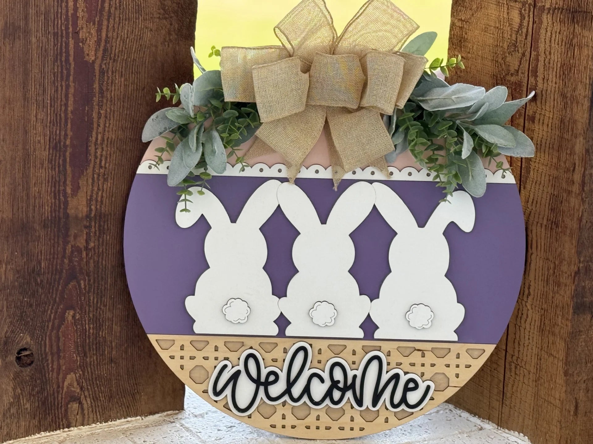A round wooden sign hangs against a rustic backdrop. It features three white bunny silhouettes with fluffy tails on a purple background, topped with greenery and a gold bow. A decorative border and the word “welcome” in cursive complete the design at the bottom.