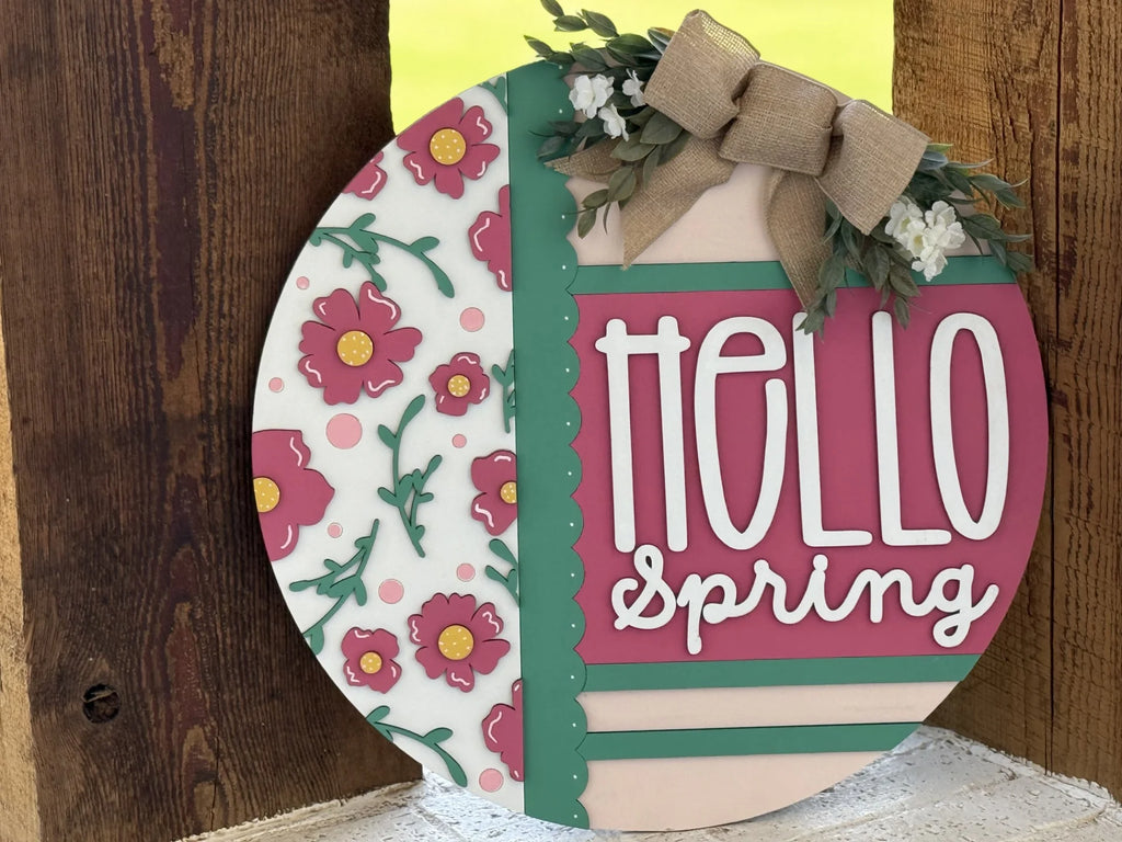 A round wooden sign says “hello spring” in playful white and pink letters. The left side has pink flowers with yellow centers. The sign is decorated with faux greenery, tiny white flowers, and a burlap bow at the top. It leans on a wooden surface.