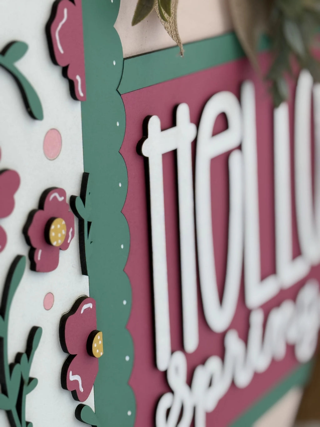 Close-up of a decorative sign with layered, cut-out paper flowers and greenery. The sign has dark pink and green colors, with the words hello spring in bold, white letters. Scalloped borders and flower embellishments add a dimensional effect.