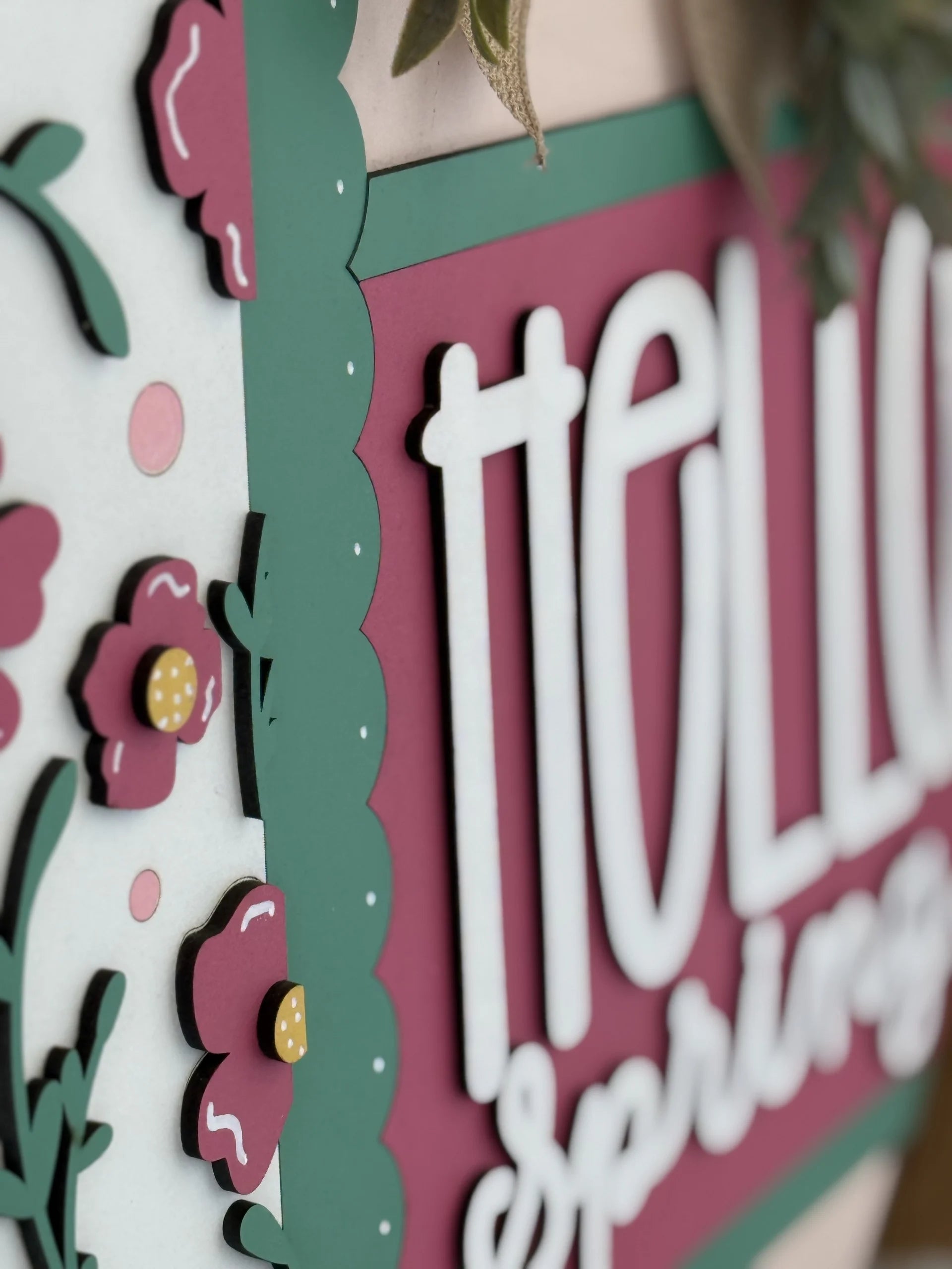 Close-up of a decorative sign with layered, cut-out paper flowers and greenery. The sign has dark pink and green colors, with the words hello spring in bold, white letters. Scalloped borders and flower embellishments add a dimensional effect.