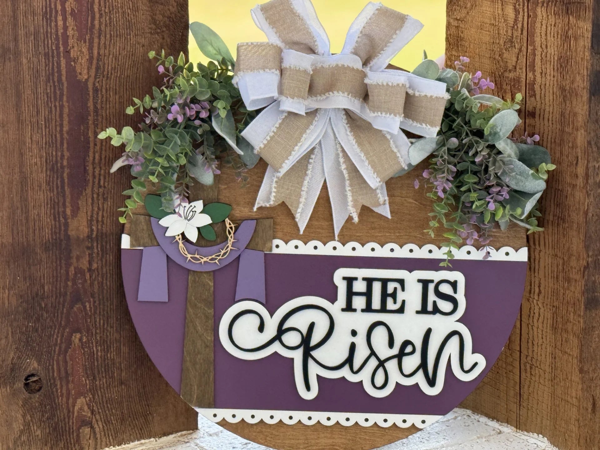 A round wooden sign decorated with greenery, purple flowers, a white and burlap bow on top, a brown cross with a white lily and crown of thorns, and the words “HE IS RISEN” in white letters on a purple background. The background is rustic wood.