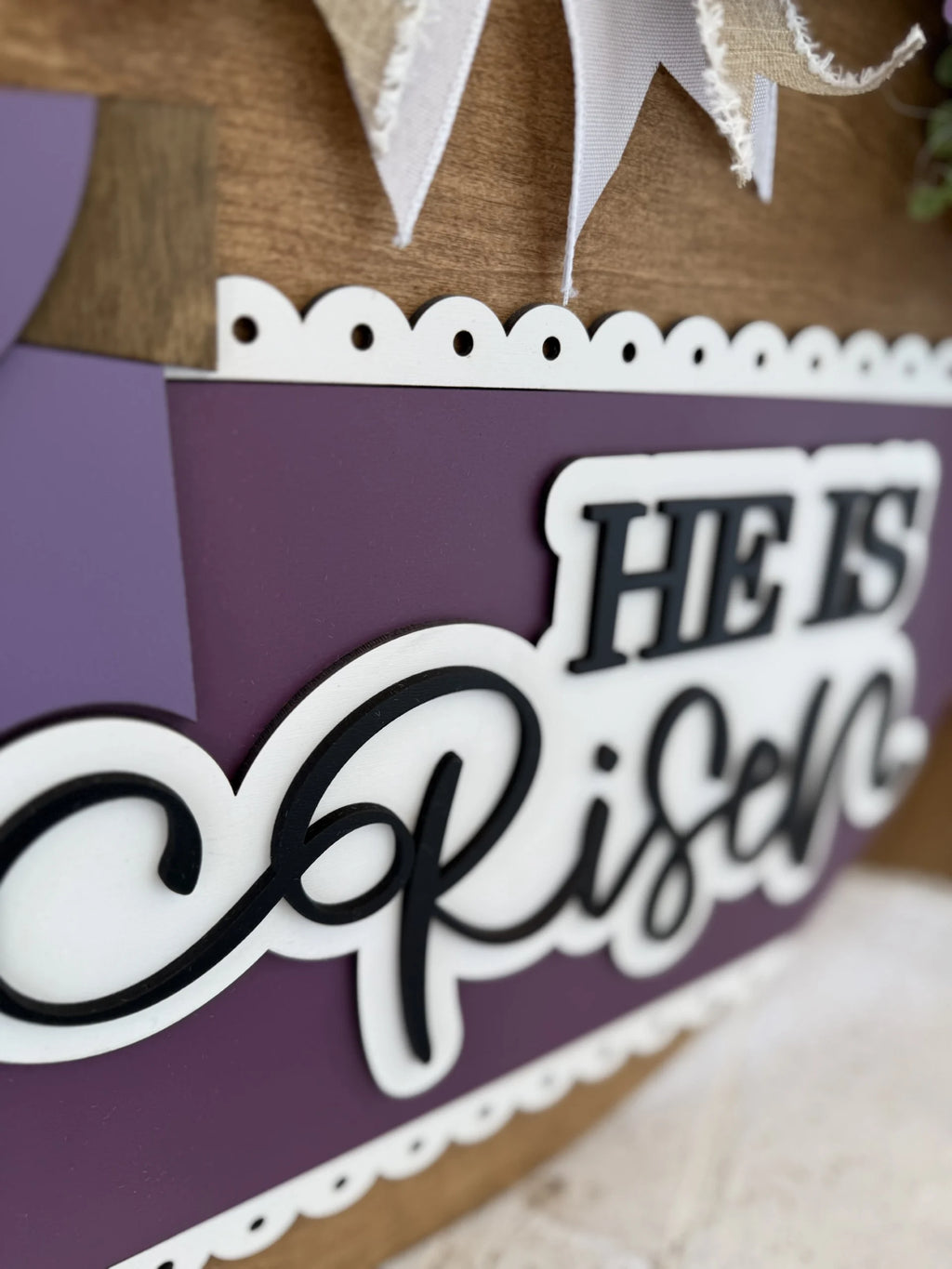 A close-up of a wooden sign with layered cutout text reading “HE IS Risen” in bold, black and white letters on a purple and white background. The top edge of the sign has scalloped trim and decorative ribbon.