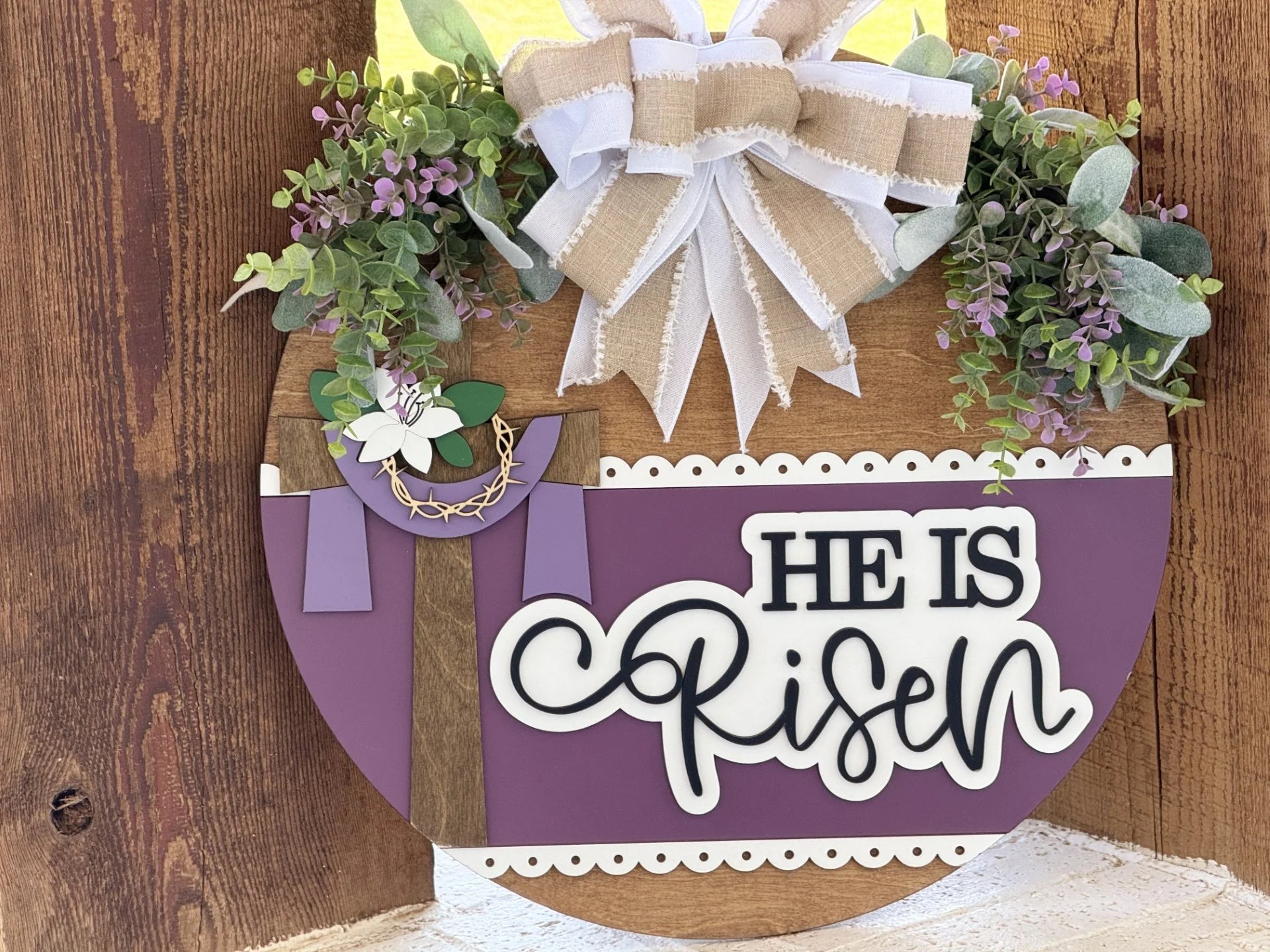 A round wooden sign decorated with faux greenery, purple flowers, and a burlap bow. On the left is a cross with a white lily and crown of thorns. On the right, bold text reads HE IS Risen on a purple background with a scalloped white border.