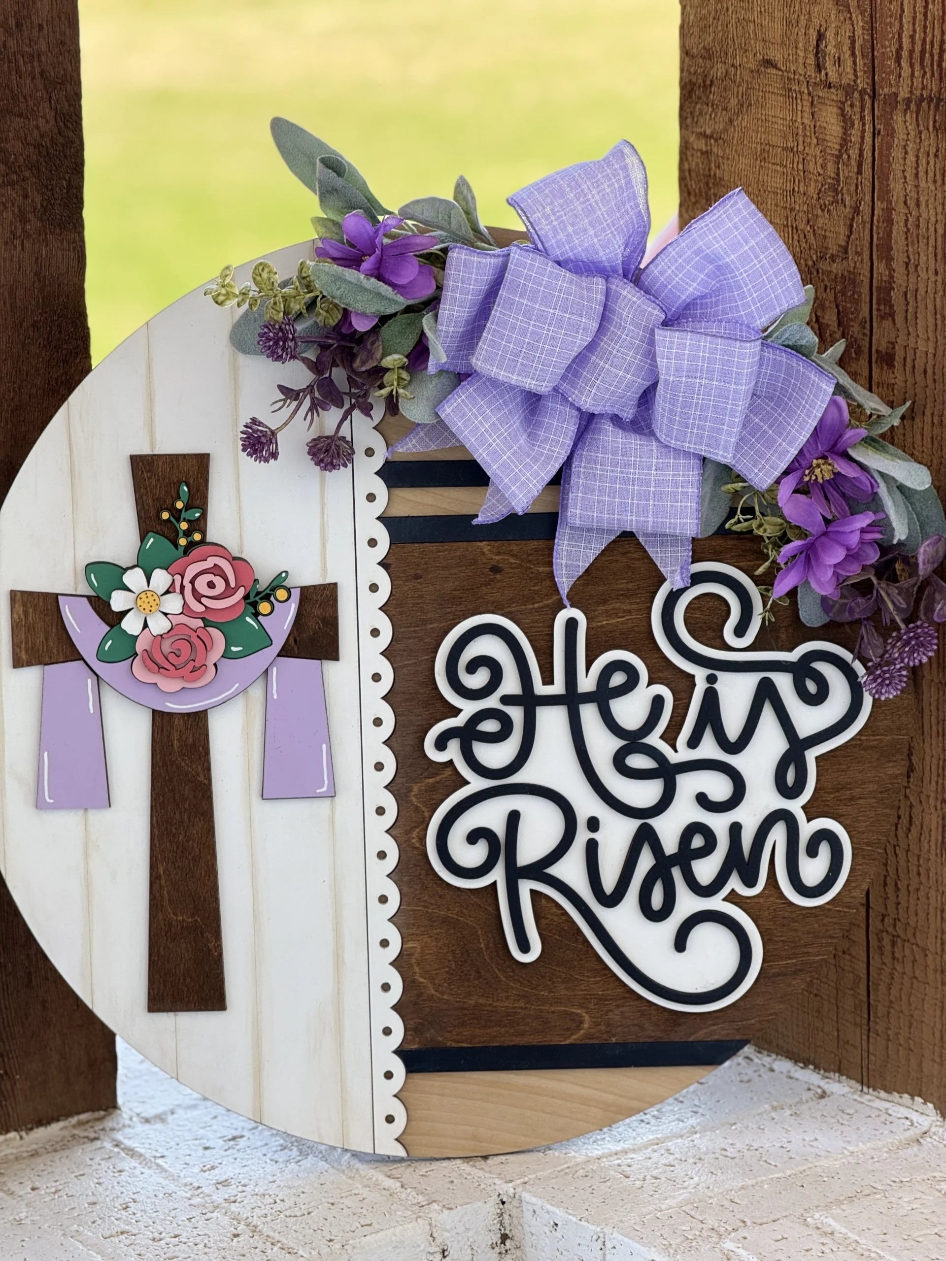A round wooden sign decorated with a cross adorned with flowers and draped with a purple cloth. The right side has “He is Risen” in script, with a large lavender bow and purple flowers at the top. The background includes stripe and scallop details.