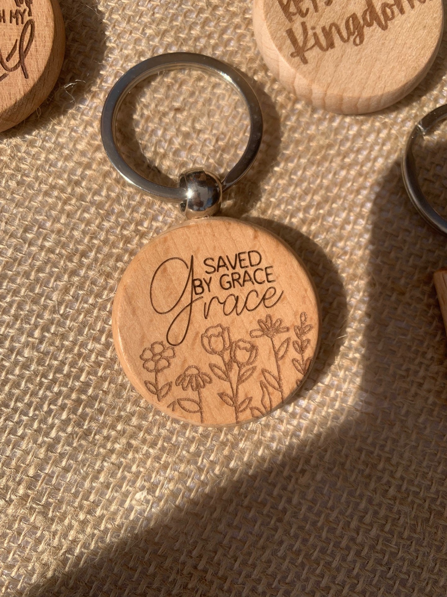 The Saved By Grace wooden round keychain showcases an elegant script with delicate flower engravings, lying on a woven burlap surface alongside another partially visible wooden item.