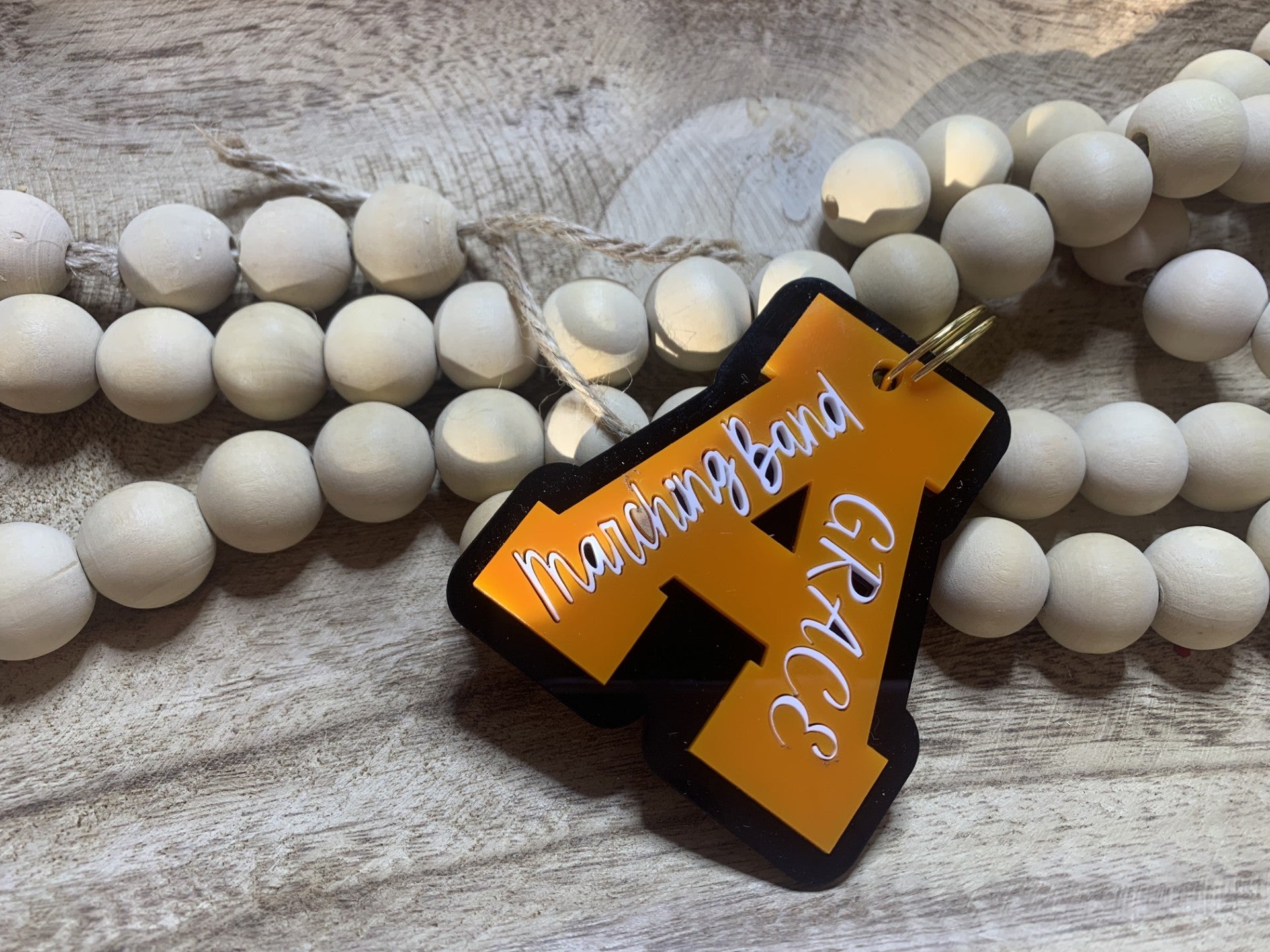 A wooden beaded garland rests on a textured wooden surface. Overlapping it is the Customized Aledo Logo Keychain, a vibrant orange and black key accessory shaped like Y with Marching Band and CRACK in white script, featuring a gold ring.