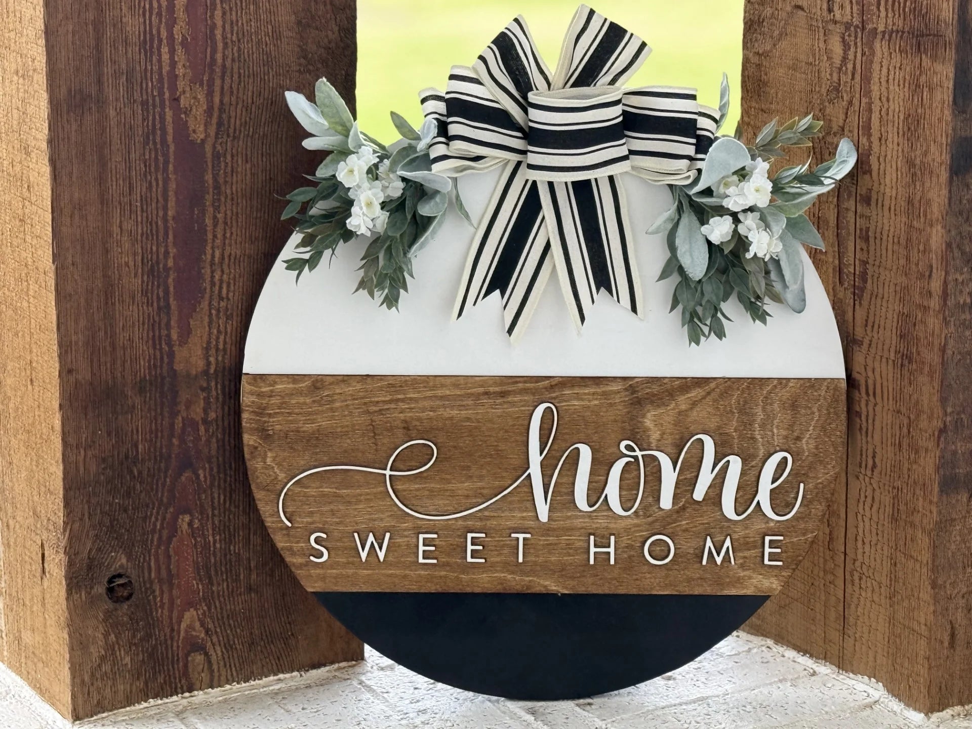 A round wooden sign with a striped black and white bow, greenery, and white flowers on top. The sign reads Home Sweet Home in white script across a brown wooden band.