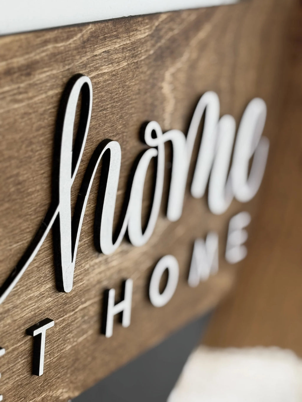 Close-up of a wooden sign with the word home written in white, cursive, raised letters. The background is brown wood with a smooth finish. The perspective is angled, focusing on the texture and lettering.