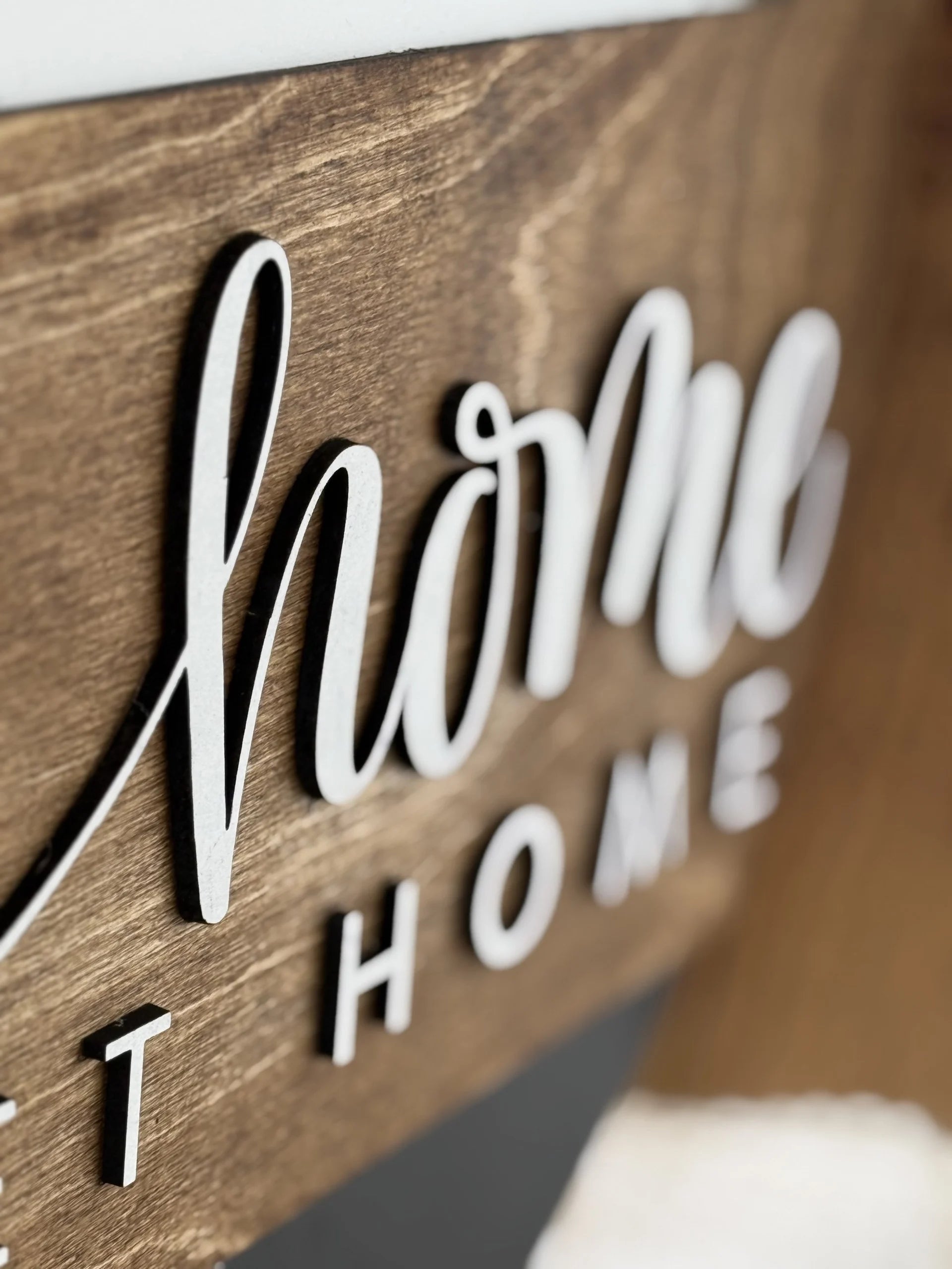 Close-up of a wooden sign with the word home written in white, cursive, raised letters. The background is brown wood with a smooth finish. The perspective is angled, focusing on the texture and lettering.