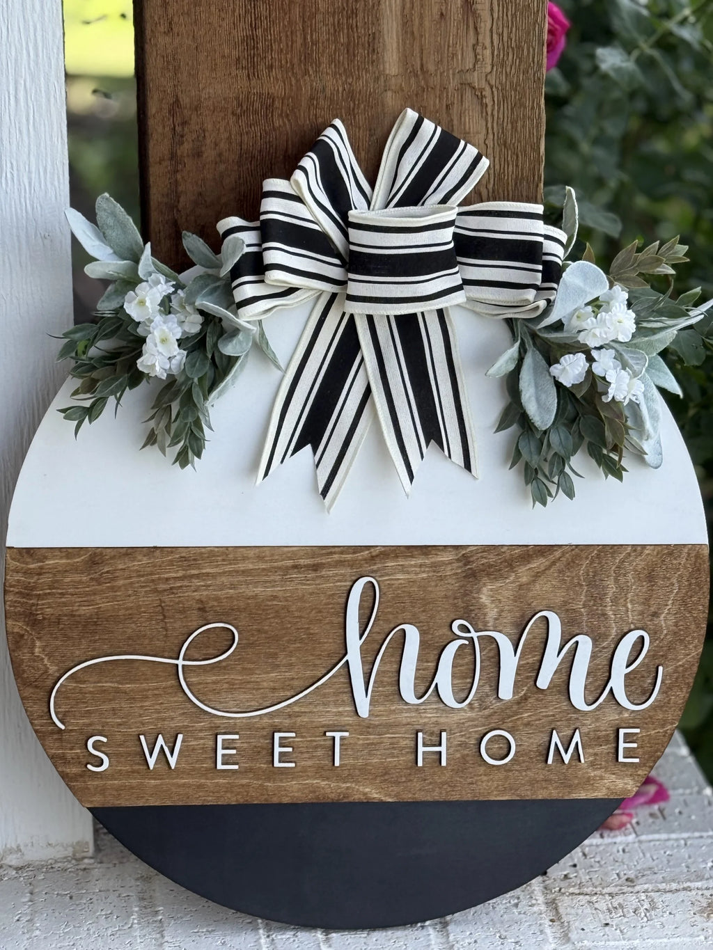 A round wooden sign with “Home Sweet Home” written in script, decorated with a black-and-white striped bow and small white flowers with greenery, hanging on a porch post.