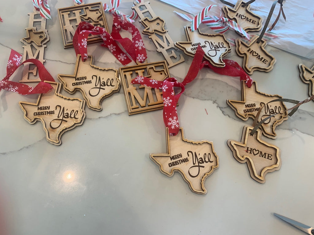 A collection of wooden Christmas ornaments shaped like the state of Texas and square plaques reading HOME are scattered on a white surface. The Texas-shaped ornaments have Merry Christmas Yall engraved and are tied with red ribbon featuring white snowflakes.