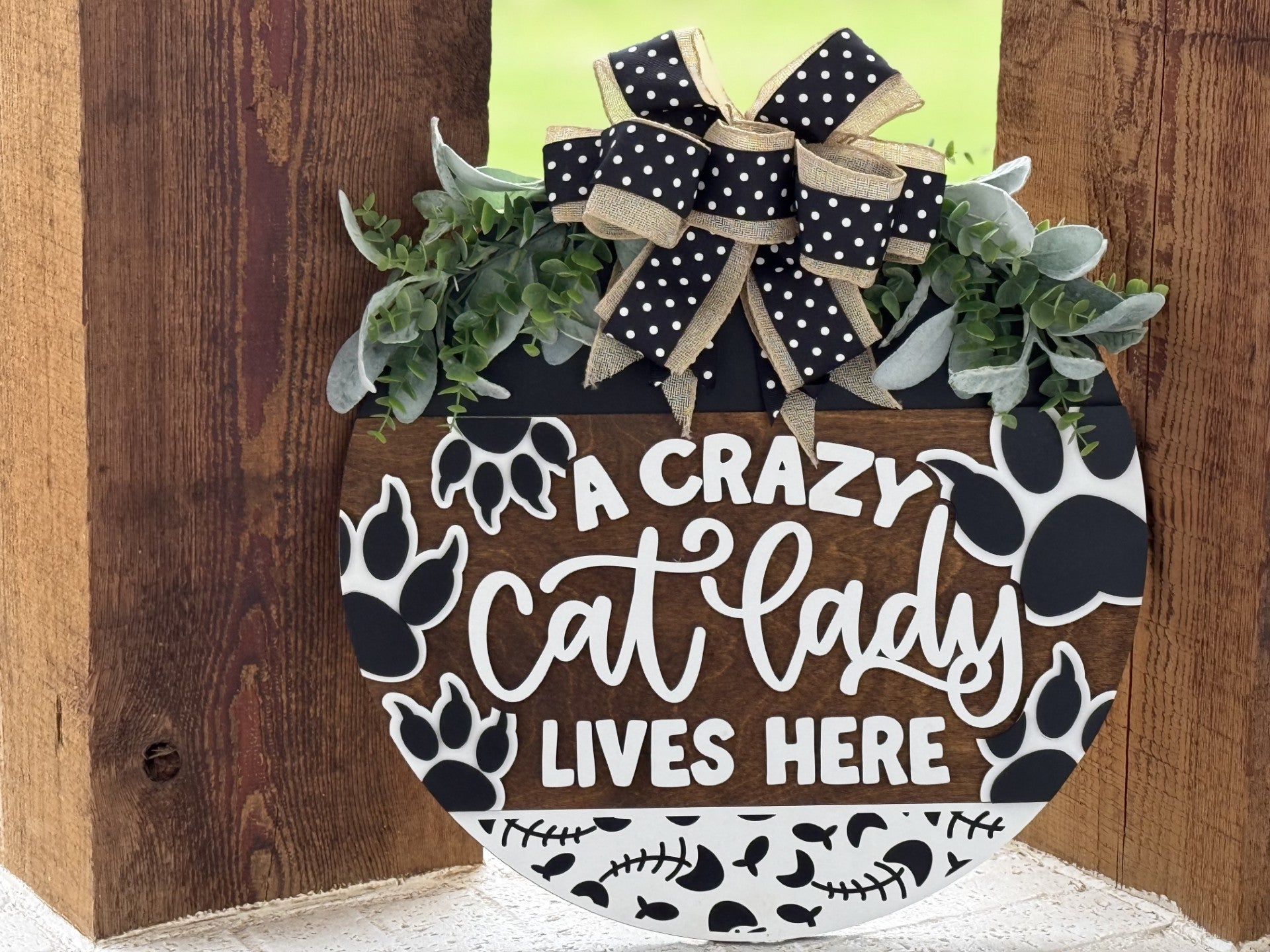 A round wooden sign with black paw prints, white bones, greenery, and a black polka-dot ribbon reads, A crazy cat lady lives here. The sign hangs on a rustic wooden background.