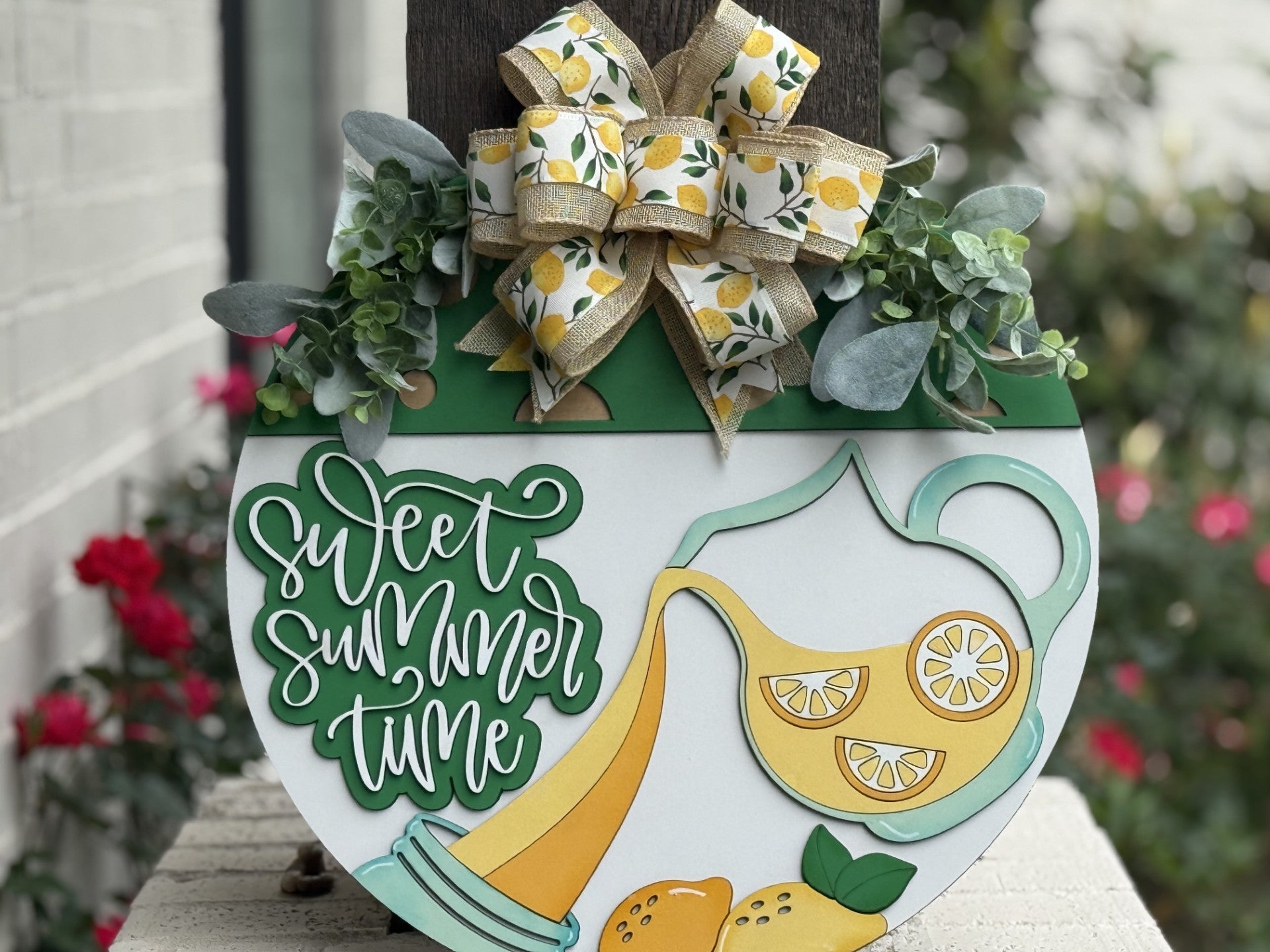 A decorative summer sign with a large bow, greenery, and lemons. The sign features a pitcher pouring lemonade and the words Sweet Summer Time in playful green lettering. Red flowers and a white brick wall are in the background.