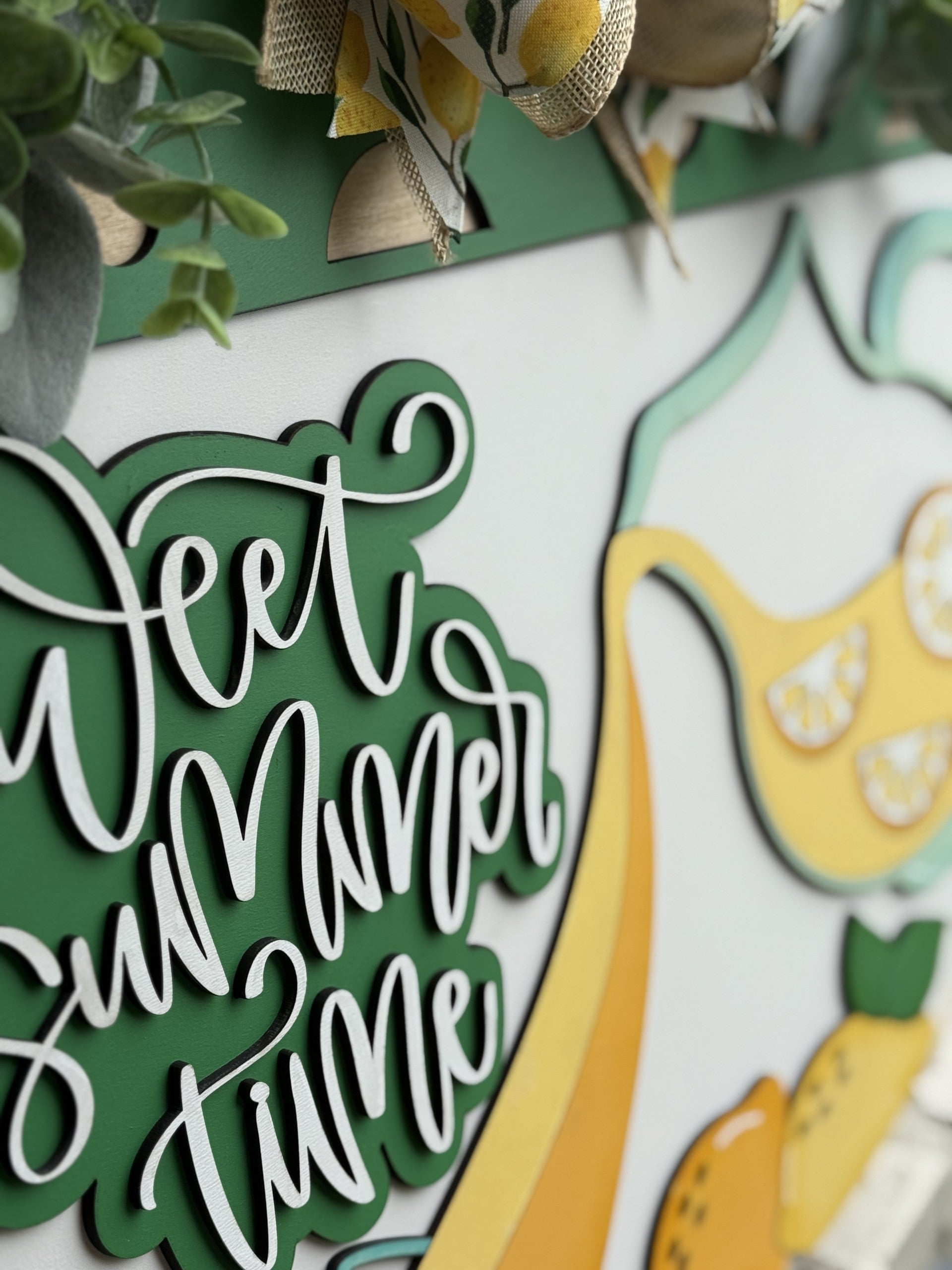 A decorative sign with the words Sweet summer time in cursive, surrounded by green leaves and lemon-themed accents. The design features yellow lemons and festive ribbon at the top.