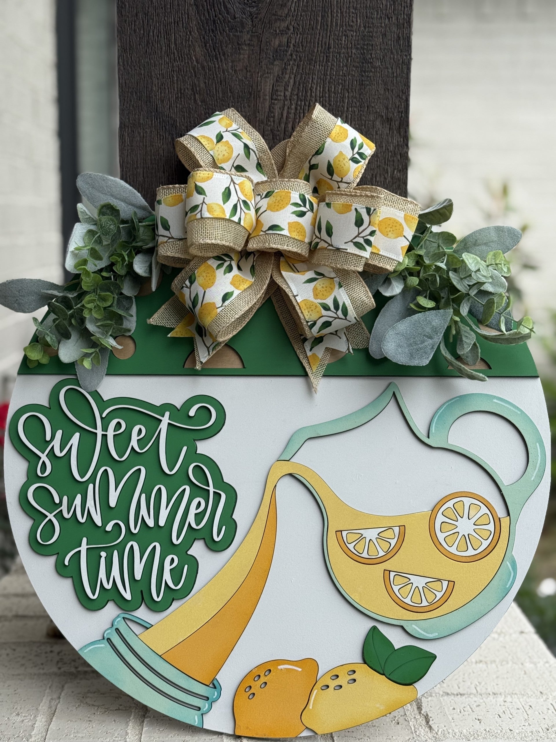A round summer-themed sign with “Sweet Summer Time” text, decorated with faux greenery, a large lemon-patterned bow, and a pitcher pouring lemonade with lemon slices and whole lemons at the bottom.