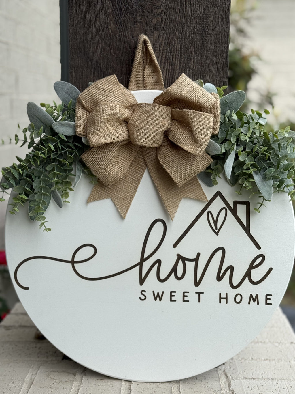 Round wooden sign with “Home Sweet Home” written in script, decorated with green leaves and a large burlap bow on top, hanging on a dark post with a white brick wall in the background.