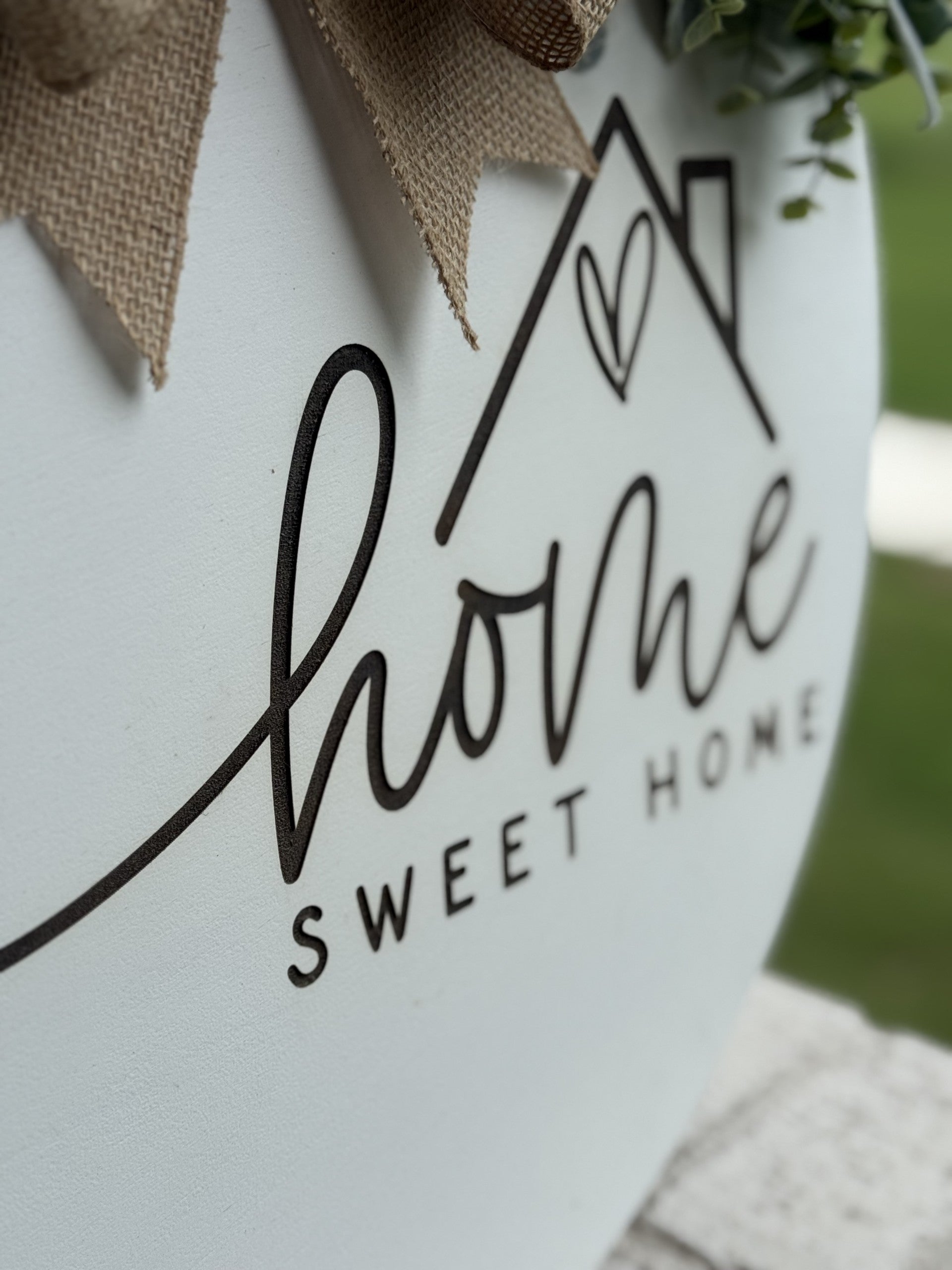 Close-up of a decorative sign with cursive black text reading Home Sweet Home and a simple house graphic with a heart, on a white background with fabric ribbon and greenery.