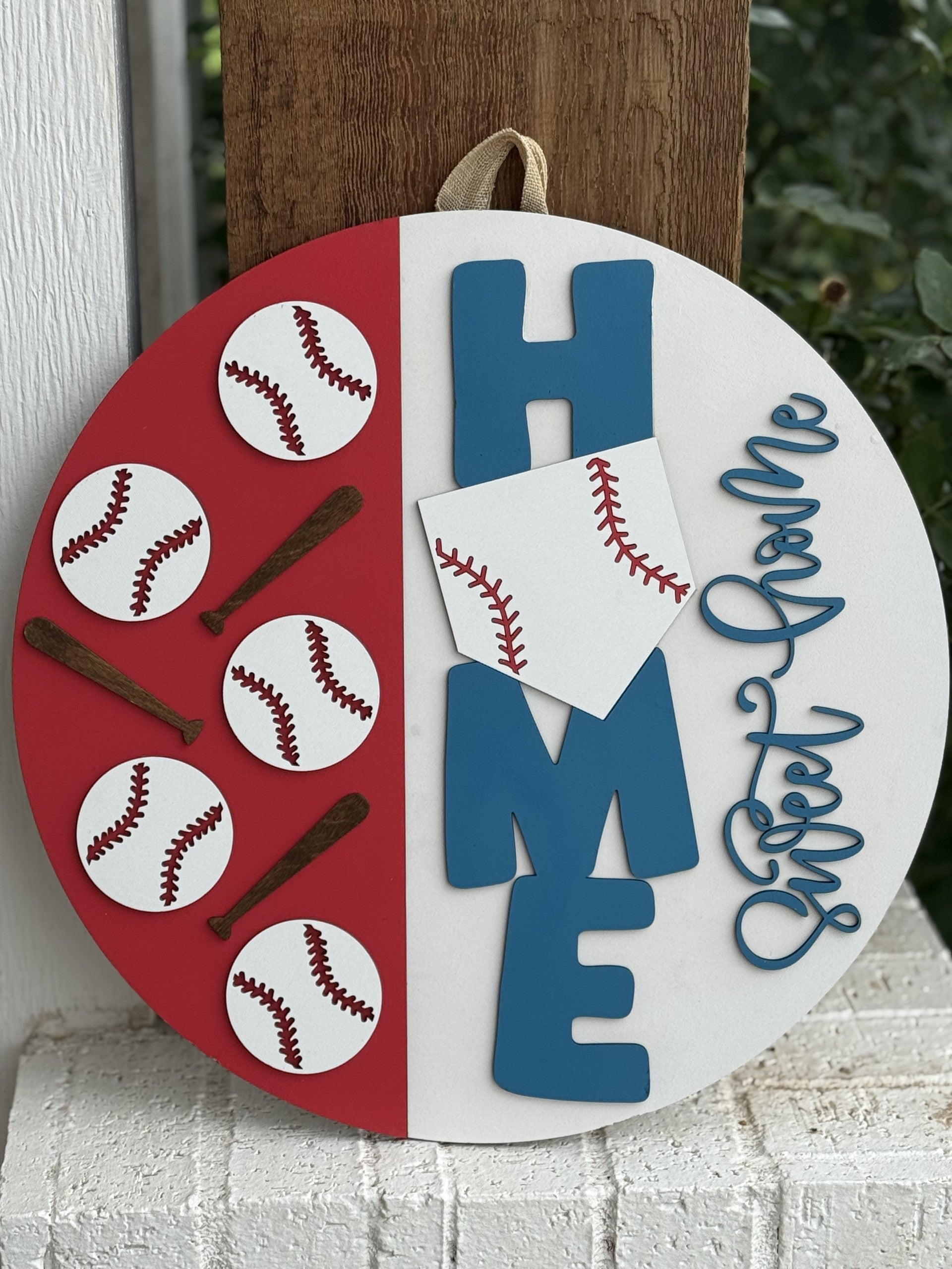 A round sign with baseballs and bats on a red half, and the word HOME in blue with a white home plate as the O. Sweet Home is written in blue script on the right side. The sign hangs by a loop at the top.