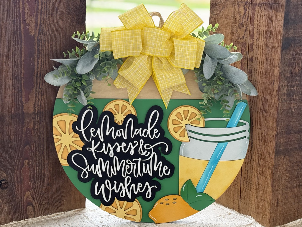 A round decorative sign with lemons, a lemonade glass, greenery, and a large yellow bow, featuring the phrase “Lemonade Kisses & Summertime Wishes” in white script lettering.