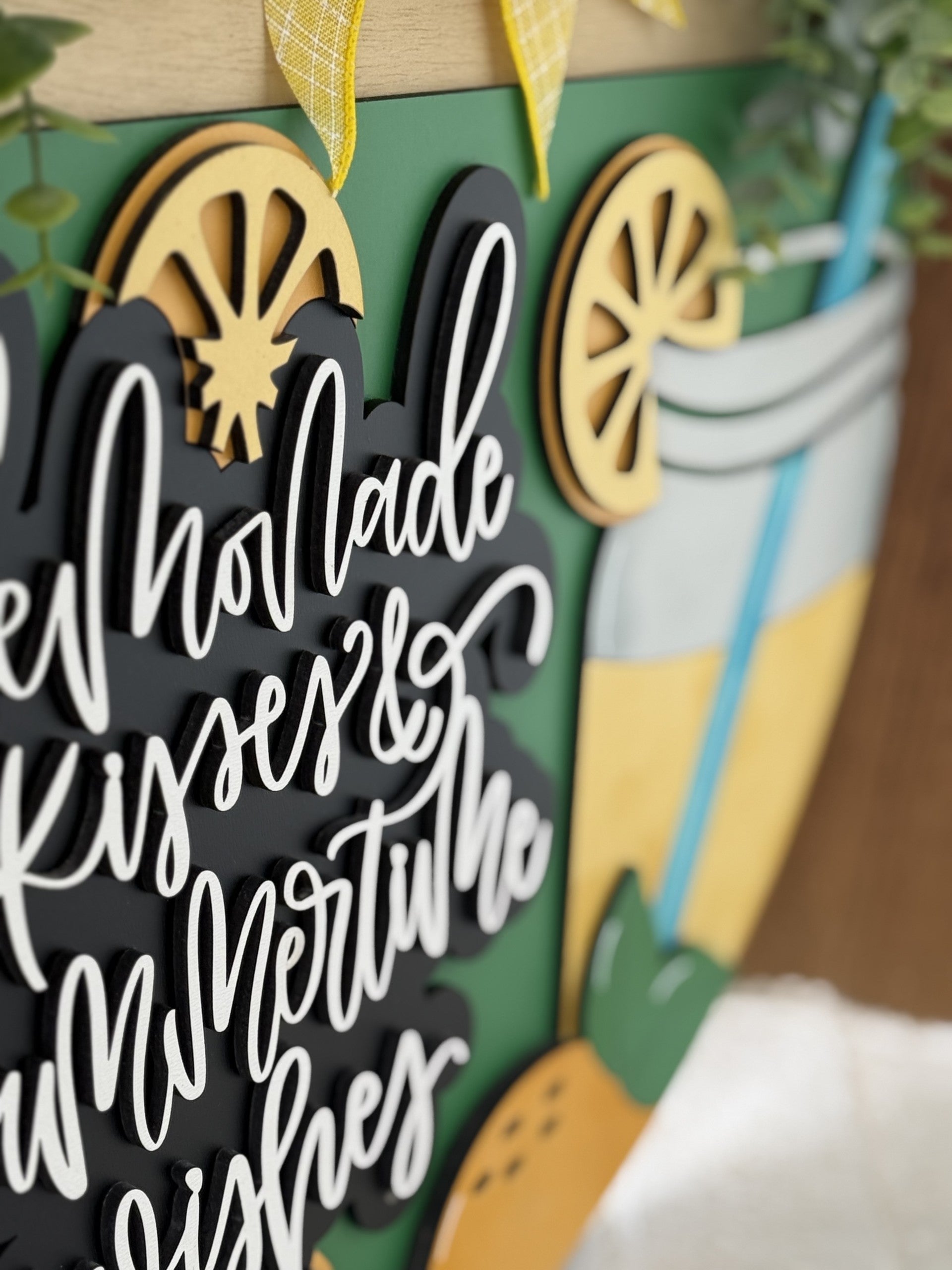 A close-up of a decorative sign with cursive white letters reading lemonade kisses & summertime wishes, featuring lemon slice cutouts and a drink illustration, accented with green leaves and yellow plaid ribbon.