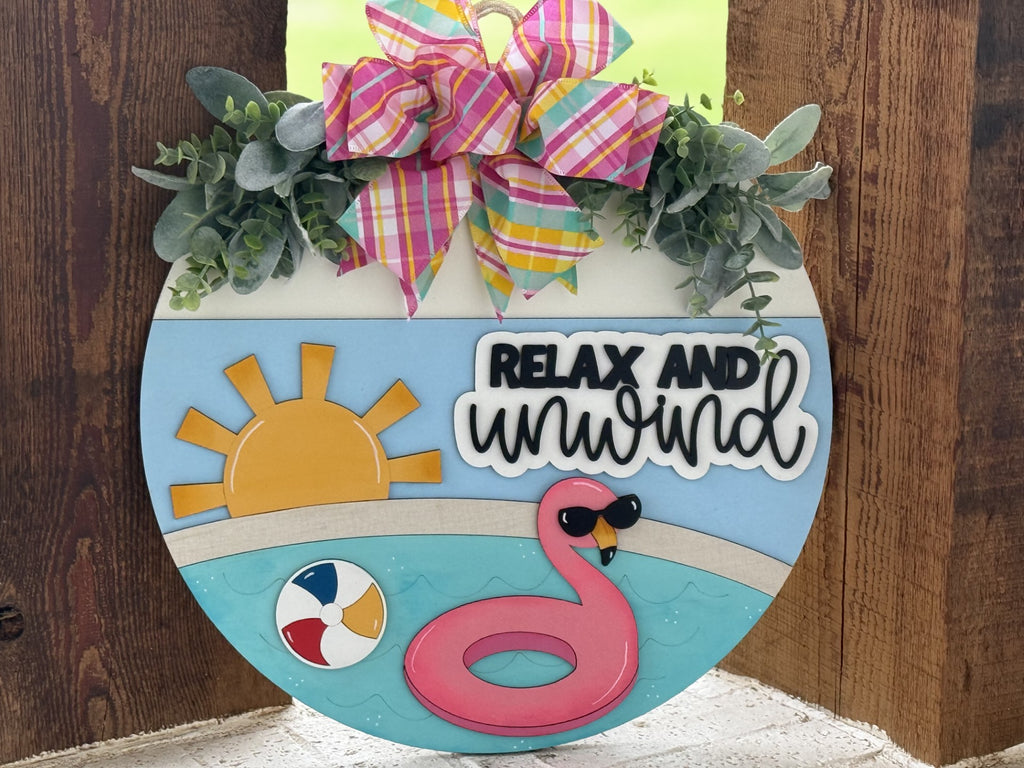 A round summer-themed door sign with faux greenery, a plaid bow, a sun, a pool with a pink flamingo float, a beach ball, and the words Relax and Unwind in black and white letters.